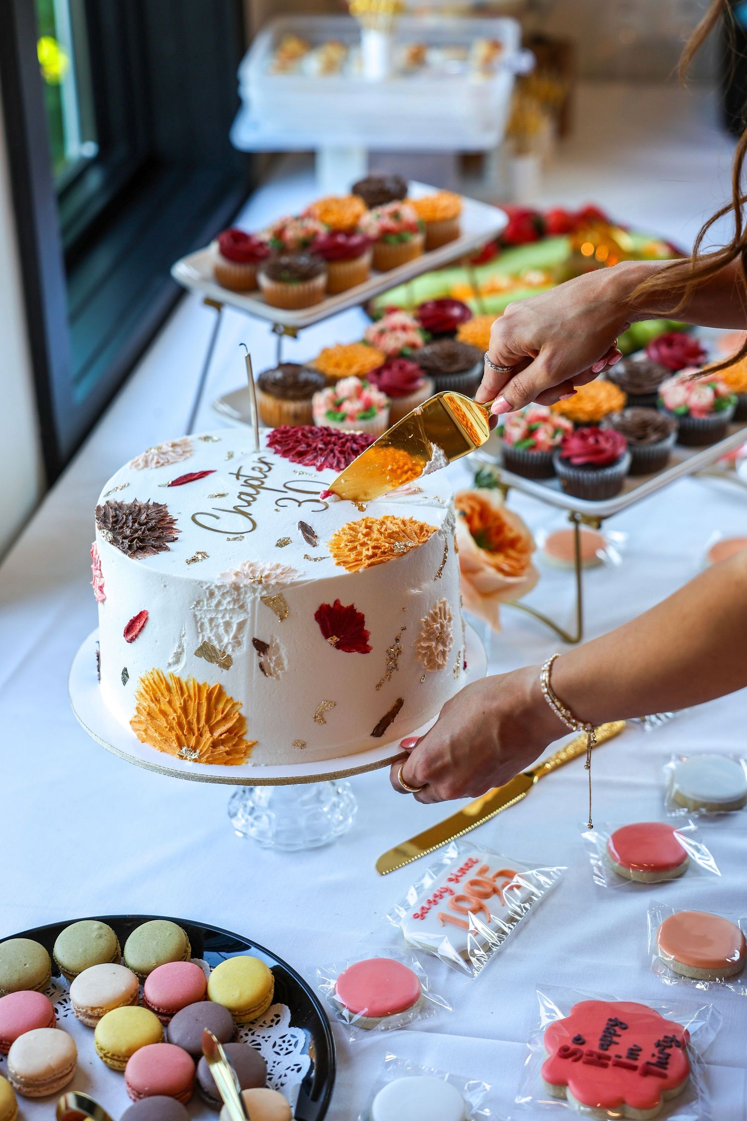 Hands cutting a floral decorated Chapter 30 birthday cake surrounded by macarons and colorful cupcakes.