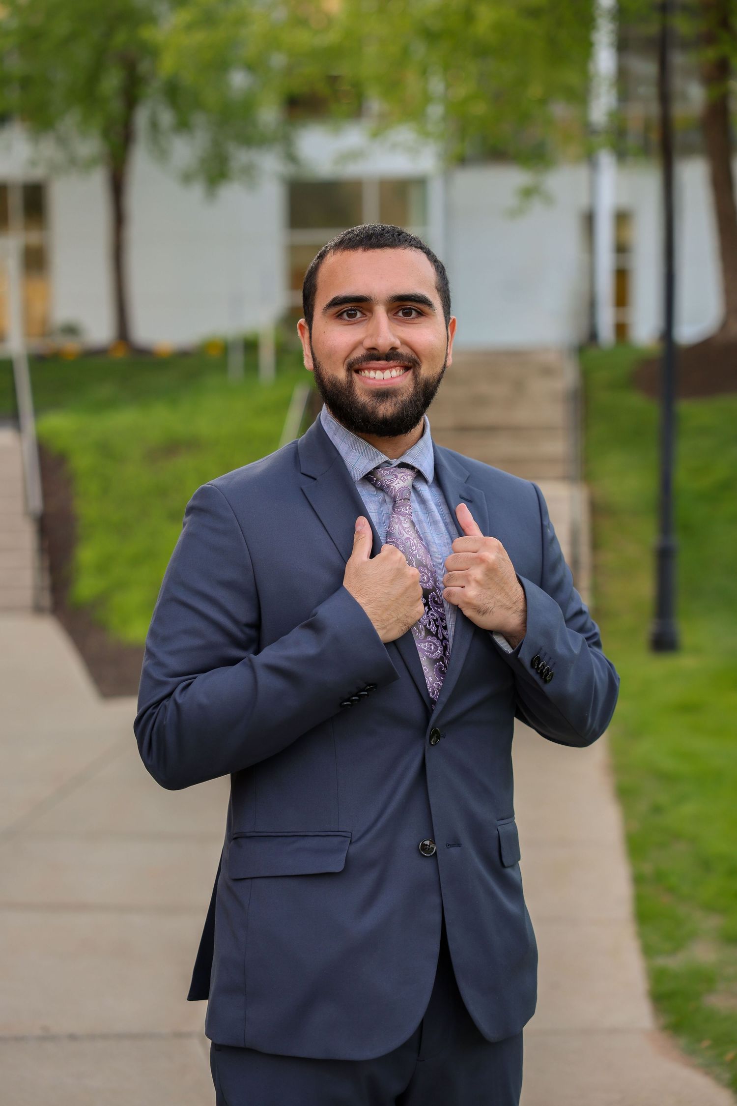 Confident bearded man in navy suit and floral tie giving thumbs up outdoors on campus.