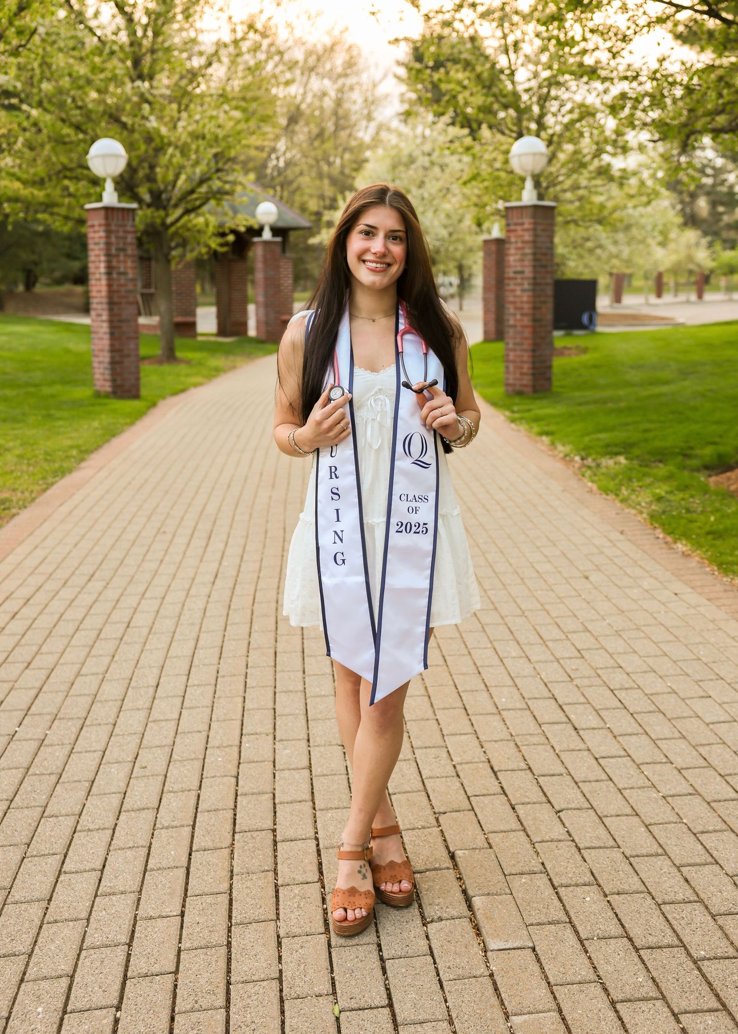 Smiling nursing graduate wearing Class of 2025 stole and white dress on brick campus pathway.