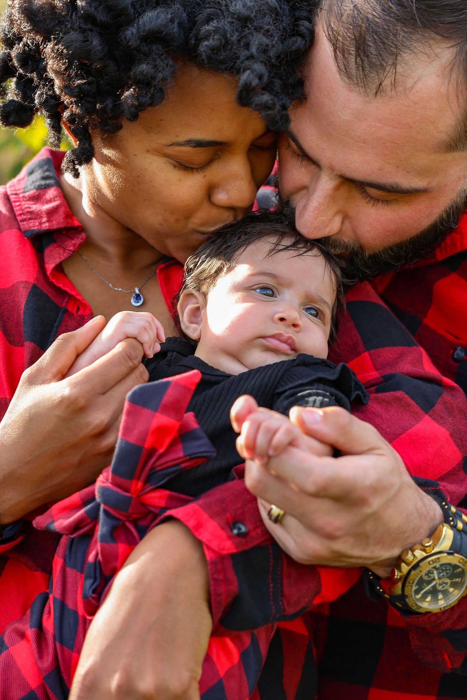 Close-up of interracial couple in red plaid shirts kissing their newborn baby on forehead in autumn setting.