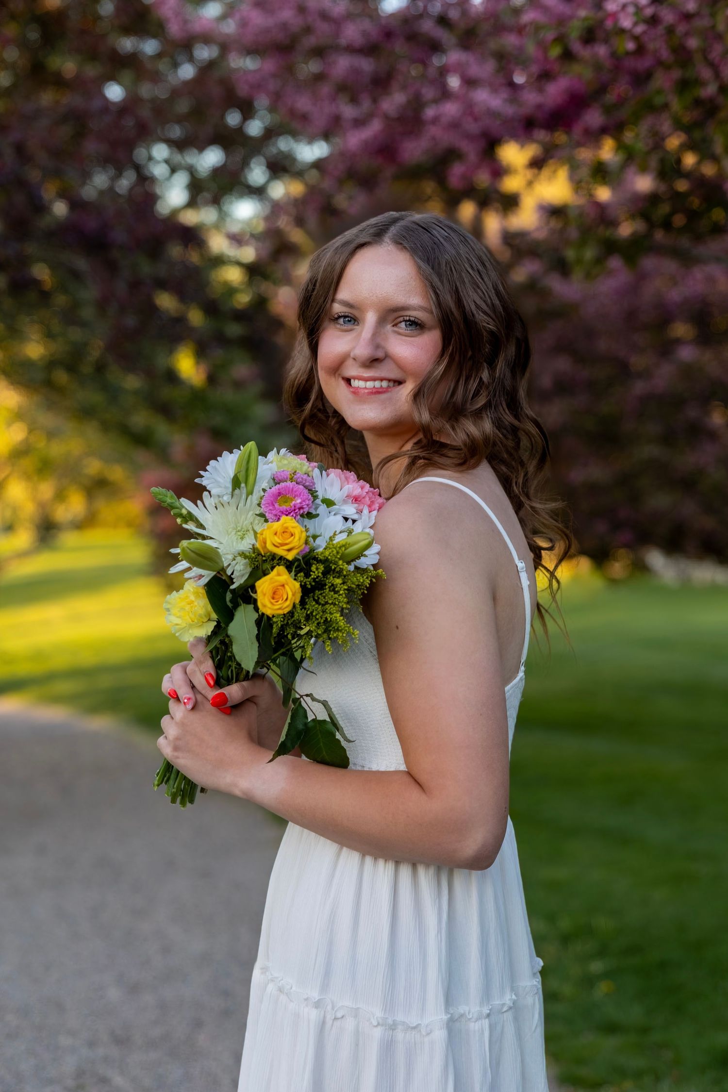 Smiling young woman in white dress holding colorful flower bouquet outdoors with blooming purple trees in background.