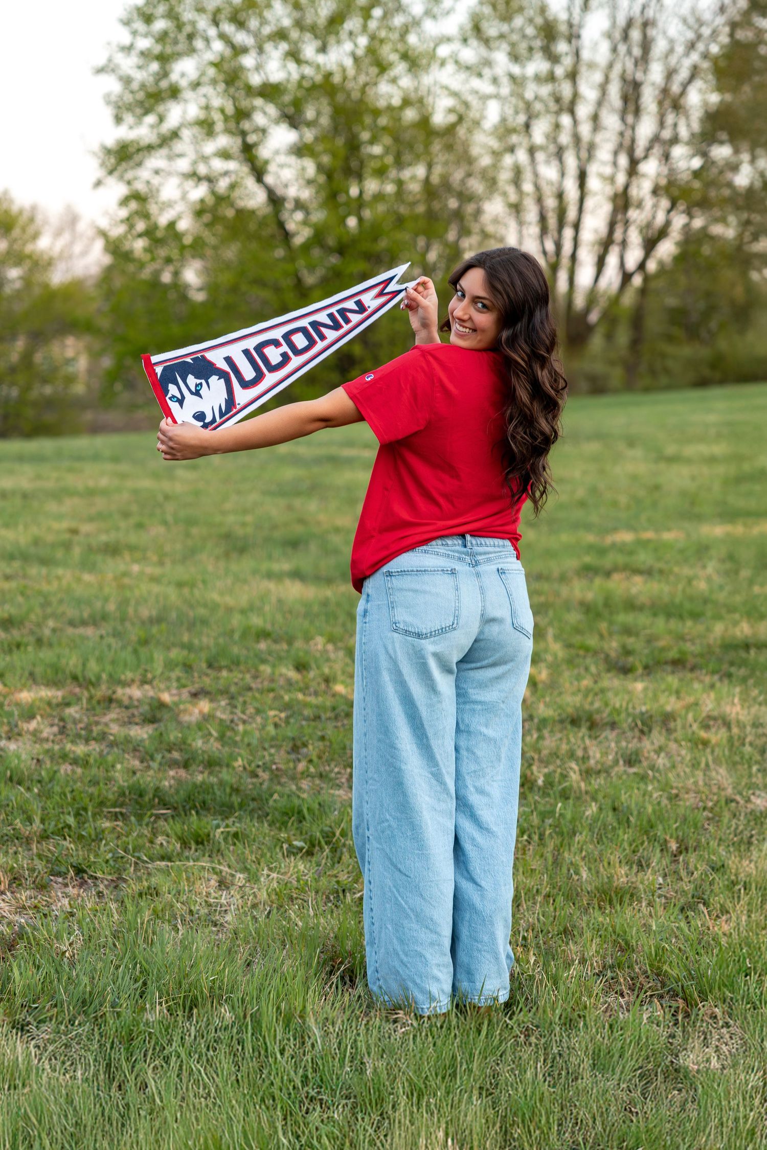 Young woman in red UConn shirt and wide-leg jeans holding university pennant in green field.