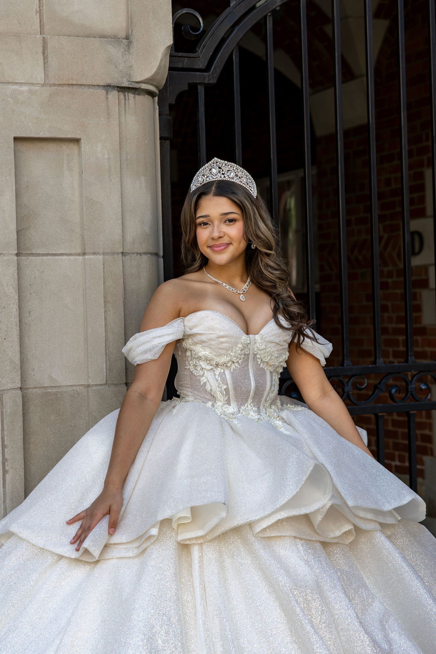 Smiling quinceañera wearing crystal tiara and white off-shoulder ball gown at iron gate.