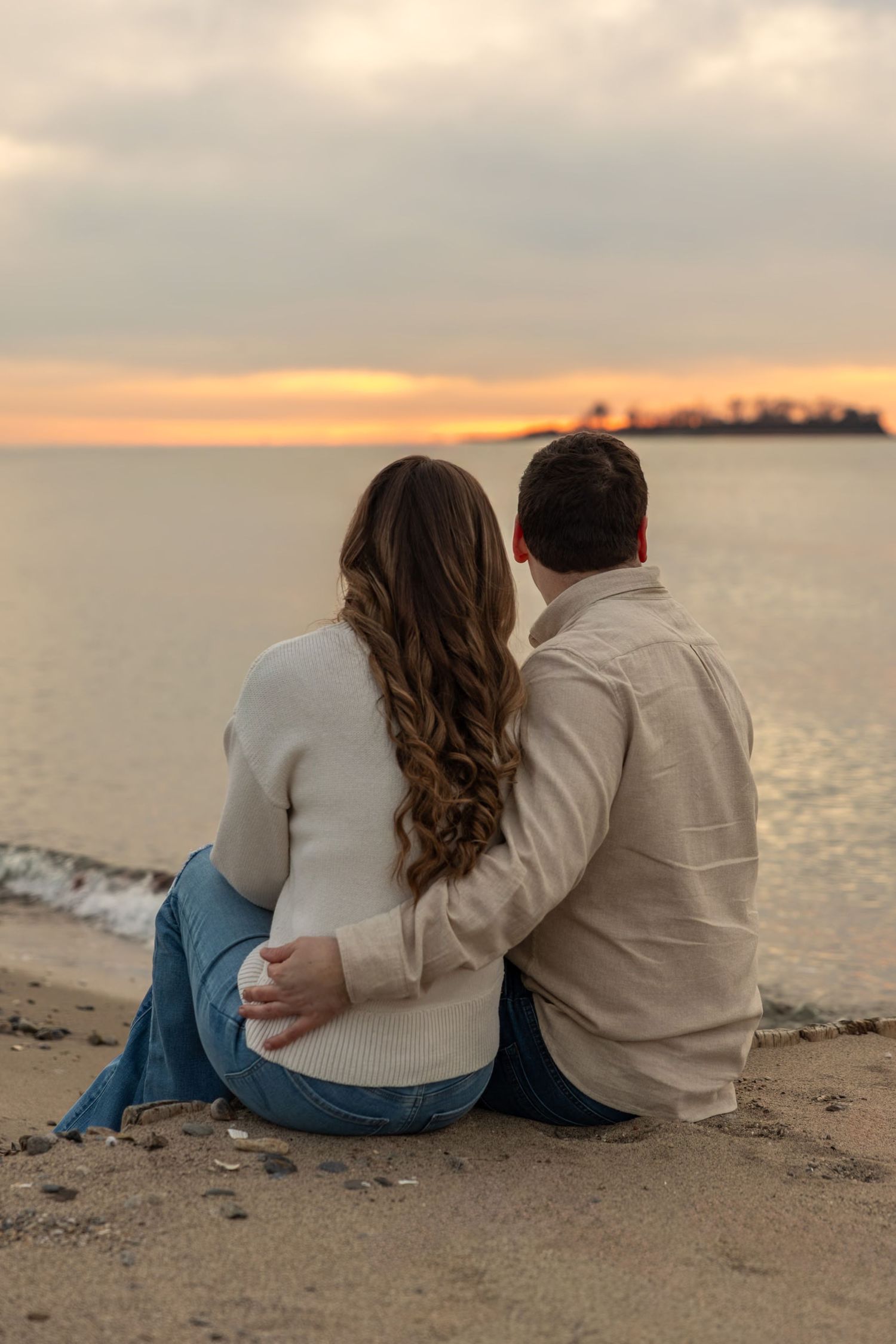 Couple sitting together on sandy beach, arms around each other watching a colorful sunset.