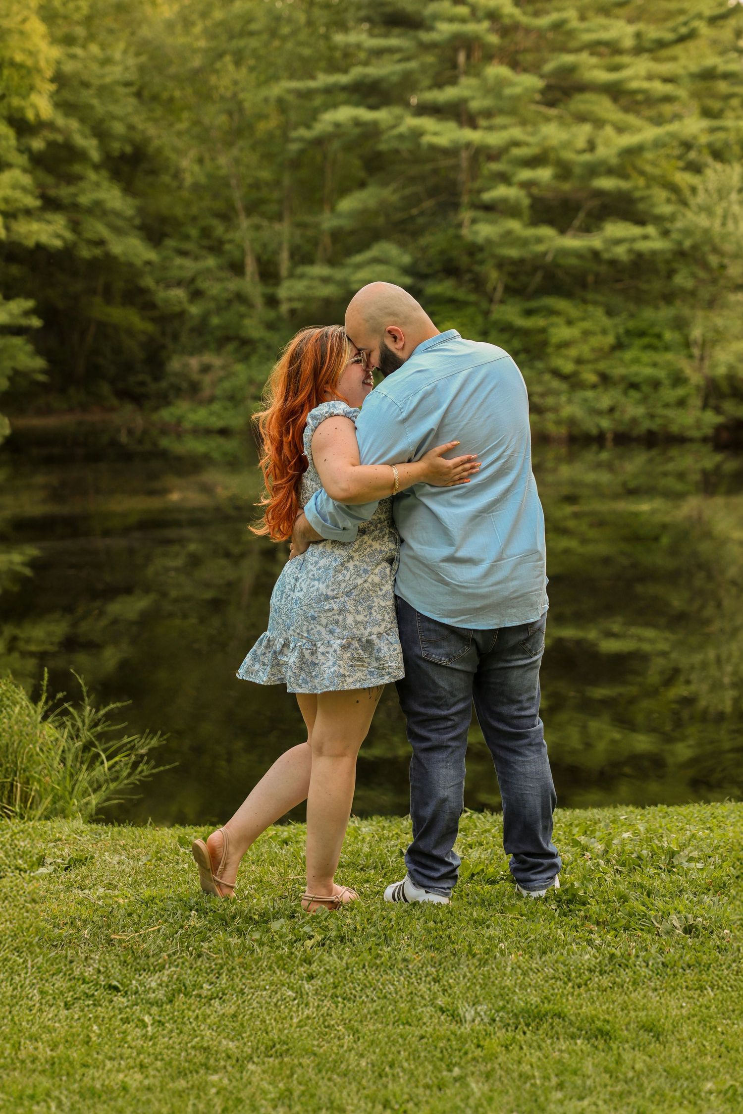 Couple embracing and sharing a kiss on lush green grass beside a serene forest pond.