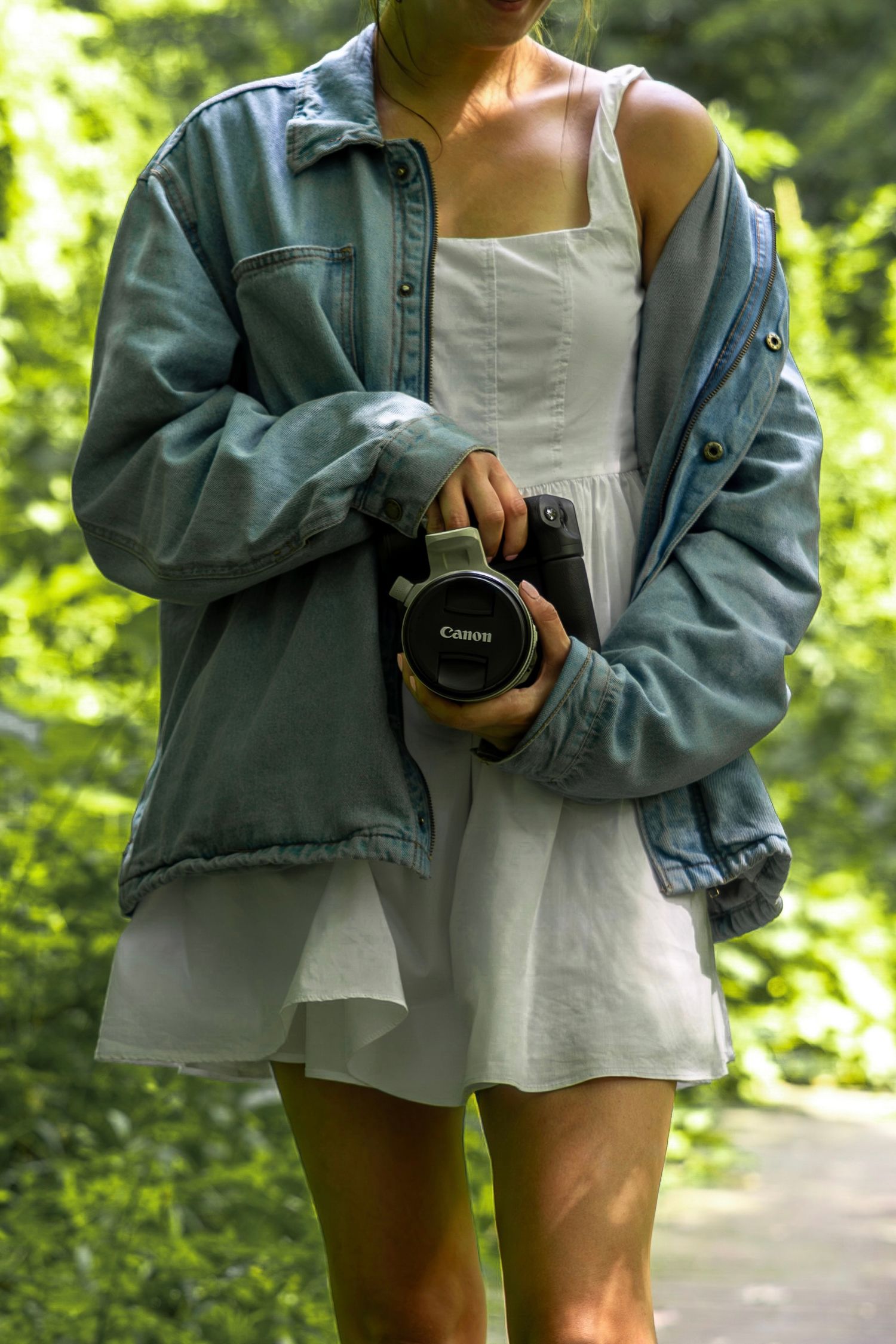 Woman in white dress and denim jacket holding a Canon camera outdoors in a lush green forest setting.