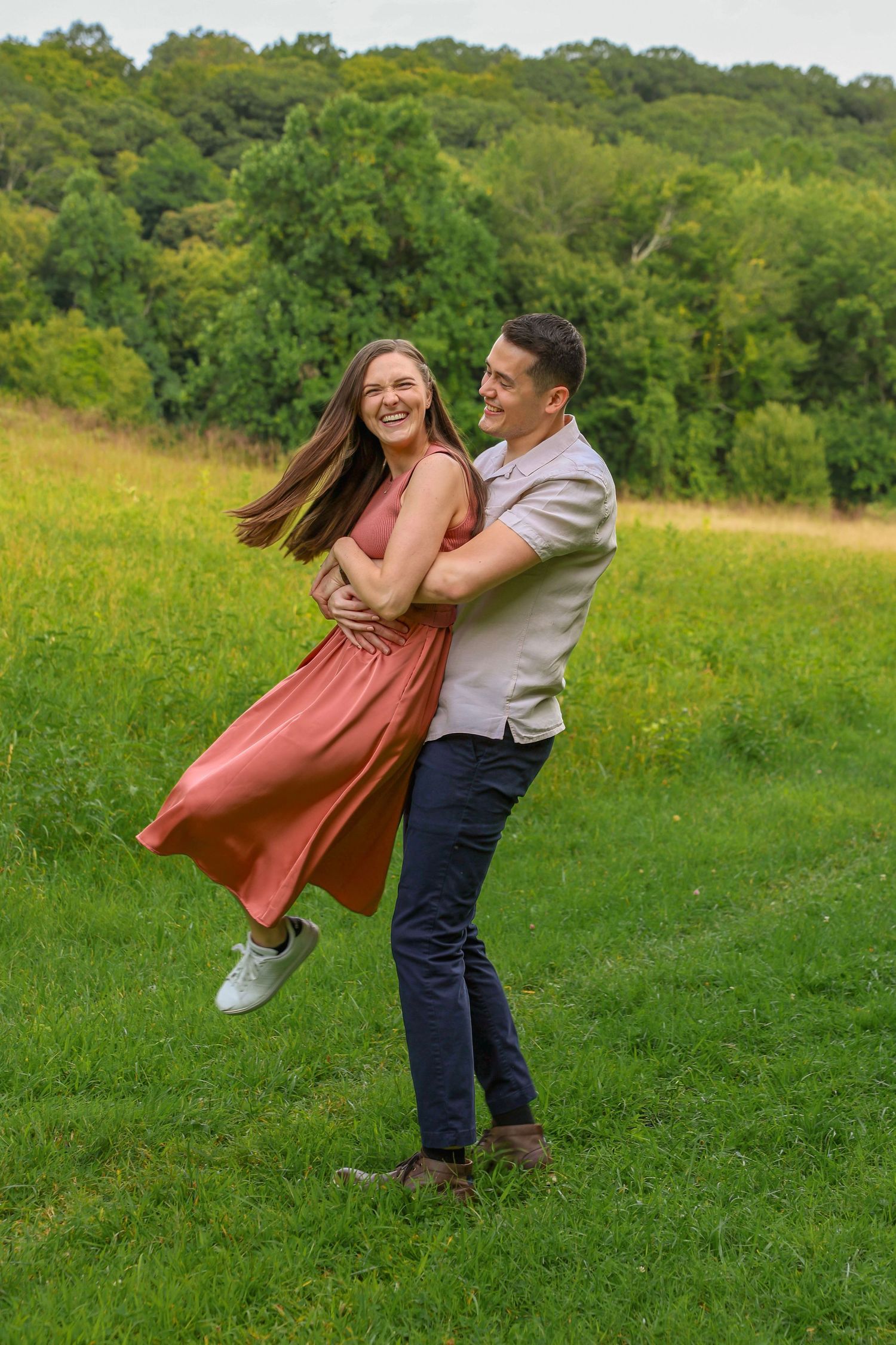 Happy couple spinning and laughing together in a lush green meadow, man lifting woman in coral dress outdoors.