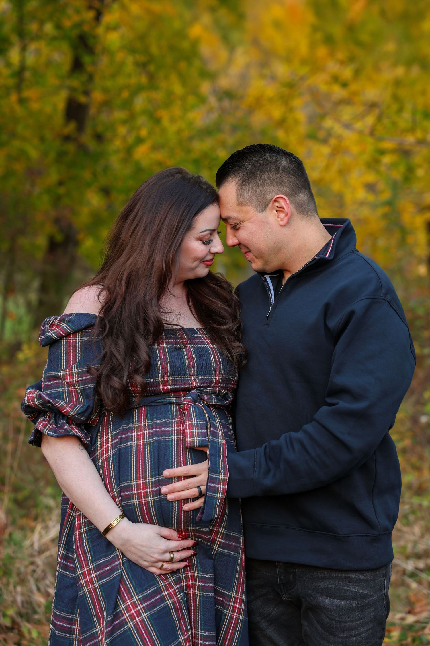 Pregnant couple touching foreheads tenderly in autumn park, woman in plaid dress cradling baby bump with golden trees behind.