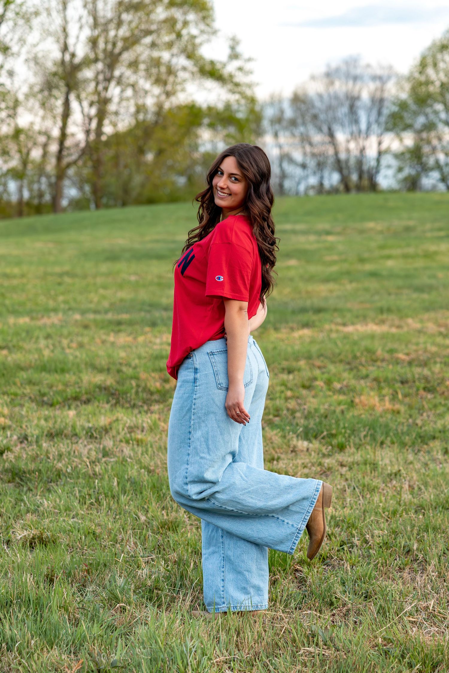Young brunette woman posing in grassy field wearing red Champion t-shirt and wide-leg light jeans.