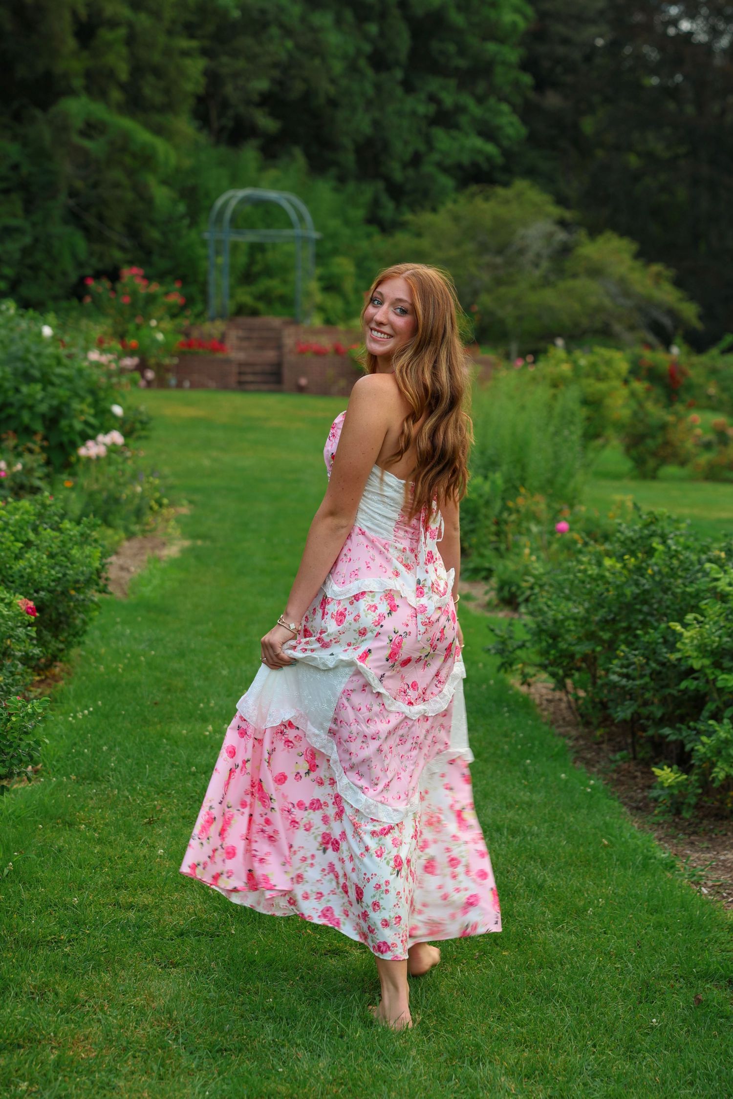 Joyful young woman in a flowing pink floral dress twirling barefoot in a lush rose garden.