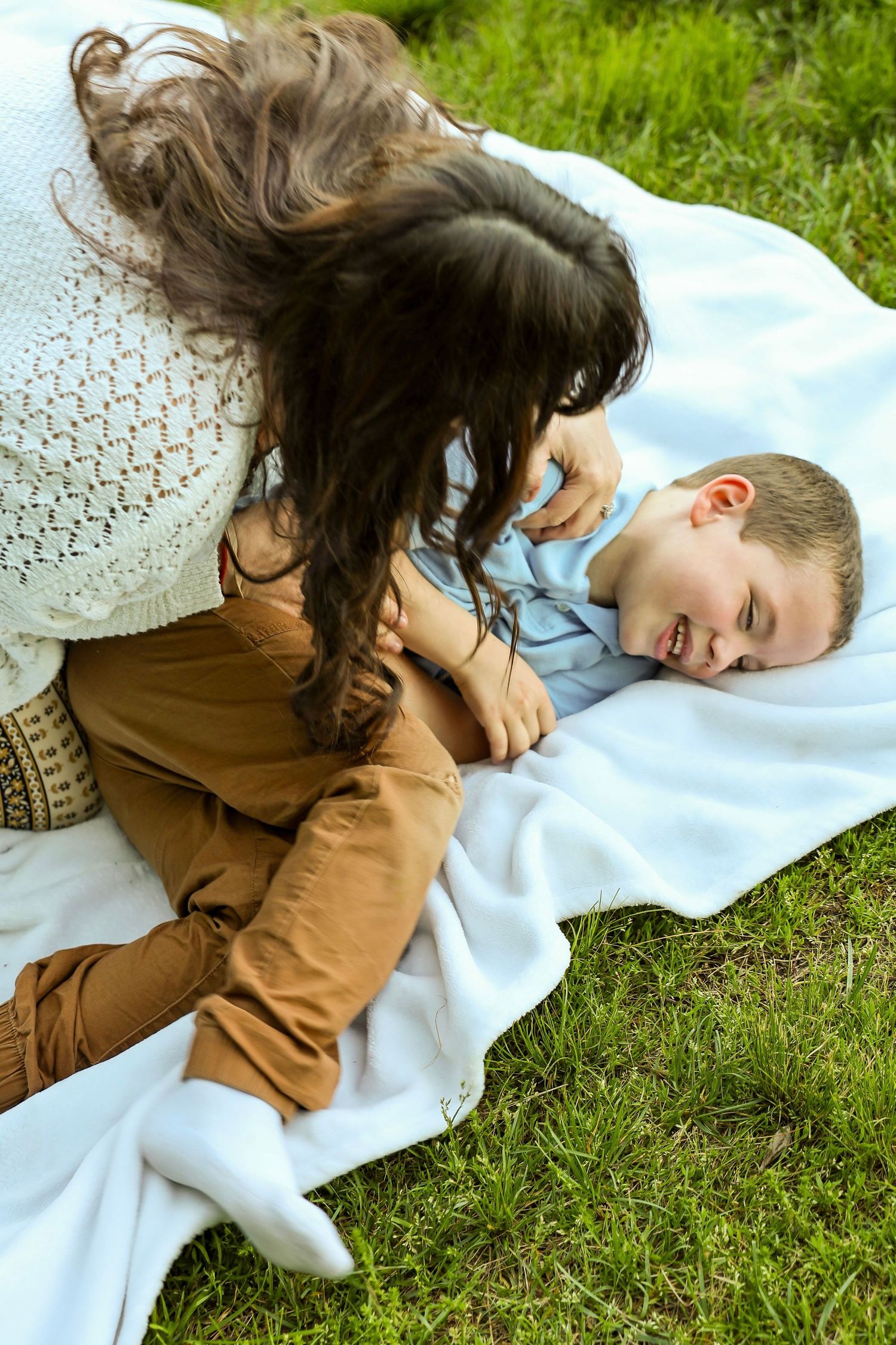 A laughing boy lies on a white blanket while his curly-haired mother playfully tickles him outdoors.