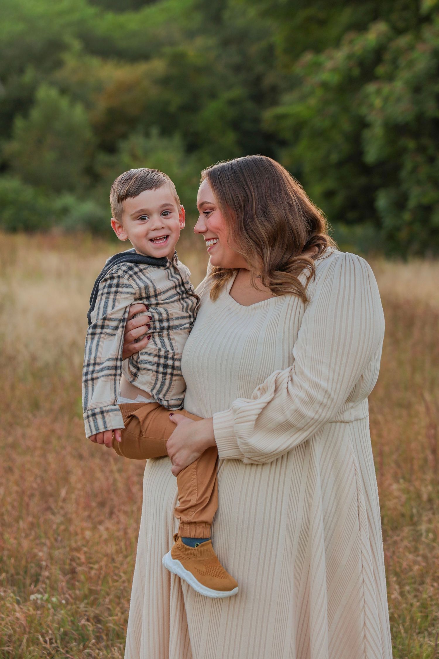 Smiling mother in cream dress holding happy toddler boy in plaid jacket in autumn field.