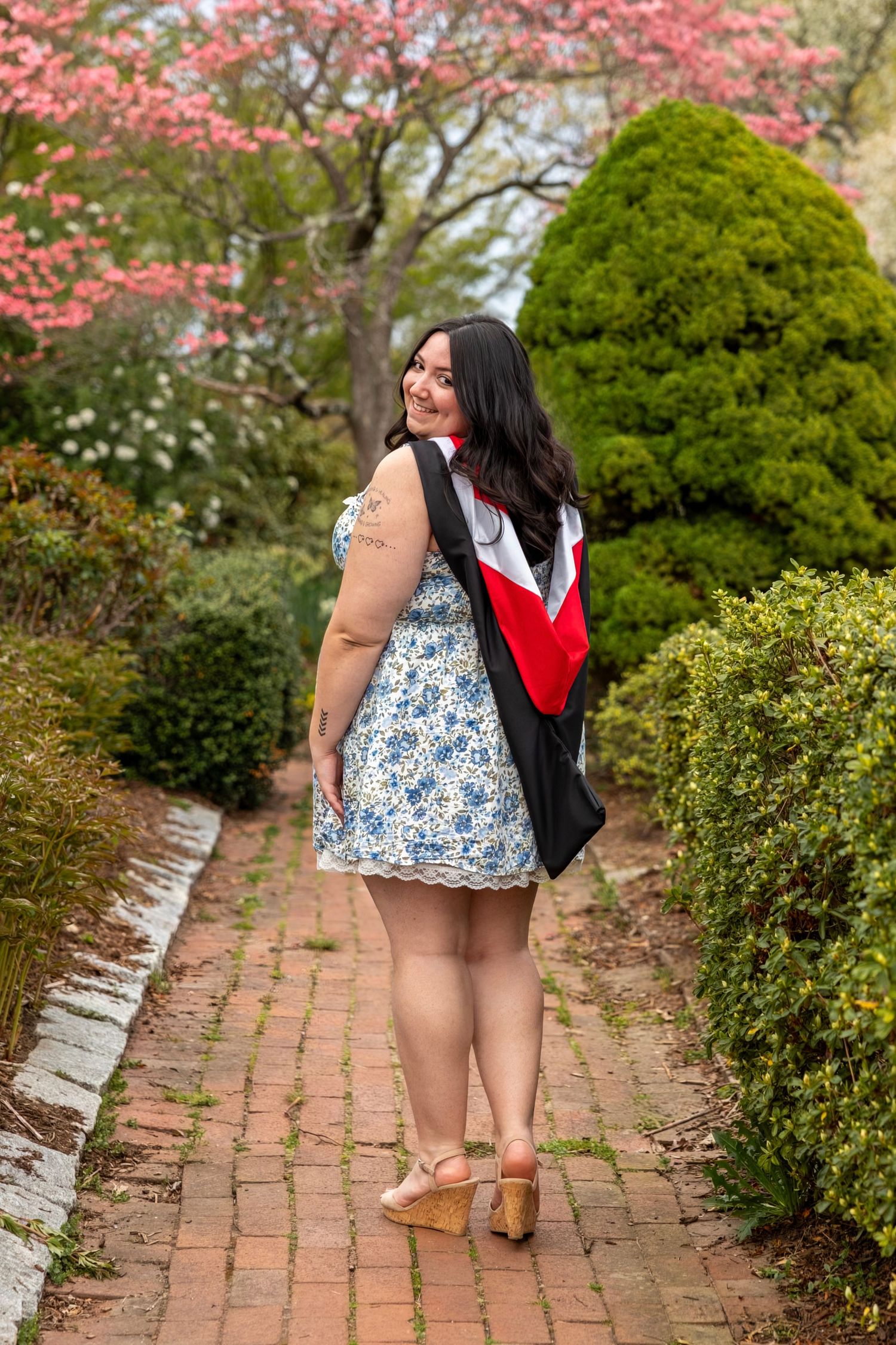Smiling graduate in floral dress with black and red academic gown poses on brick path in spring garden.