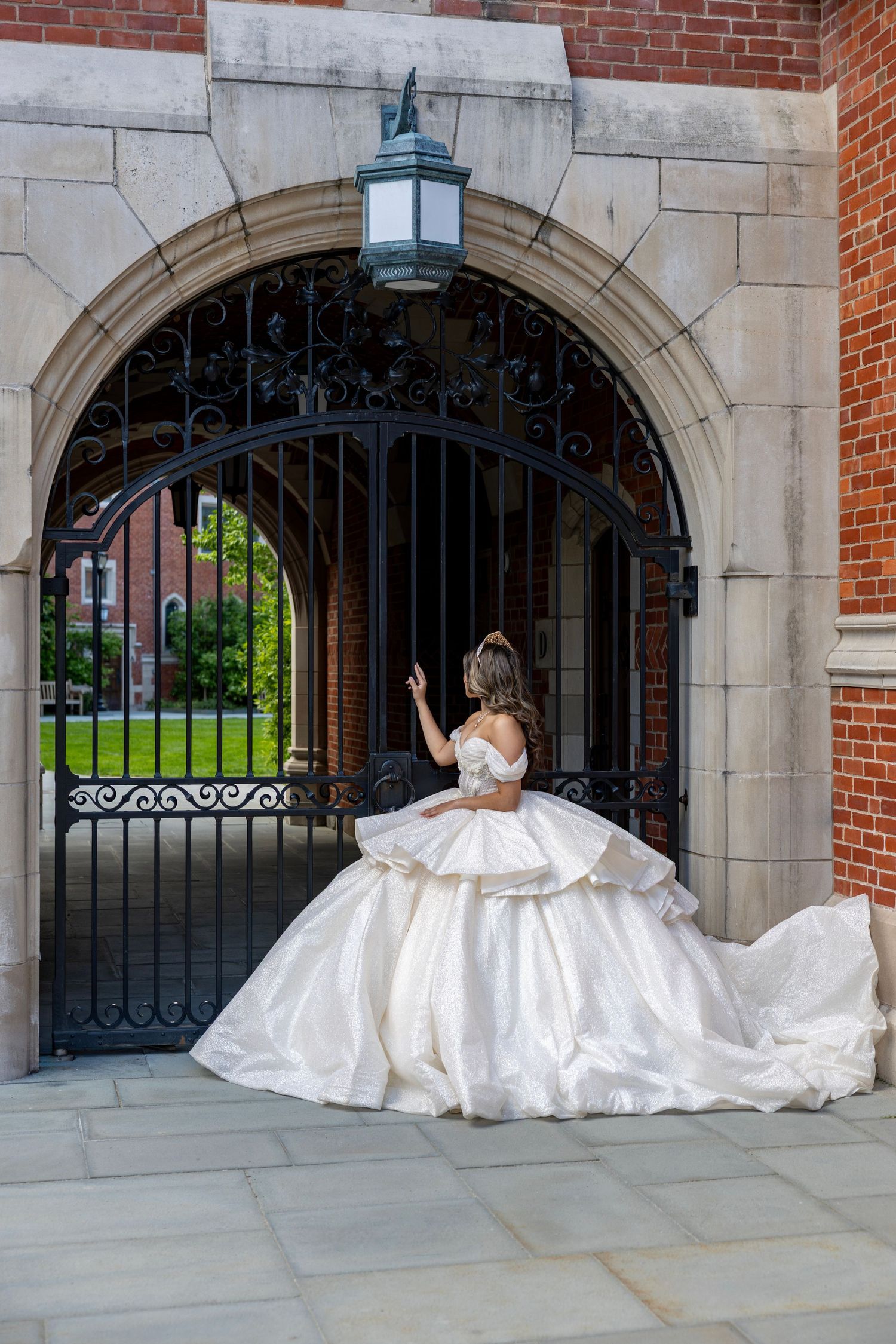 Bride in voluminous white ball gown standing at ornate iron gate archway.