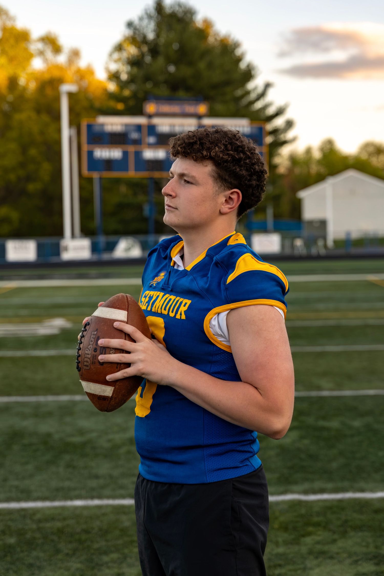 Seymour High School football player number 3 in blue jersey holding football at sunset.