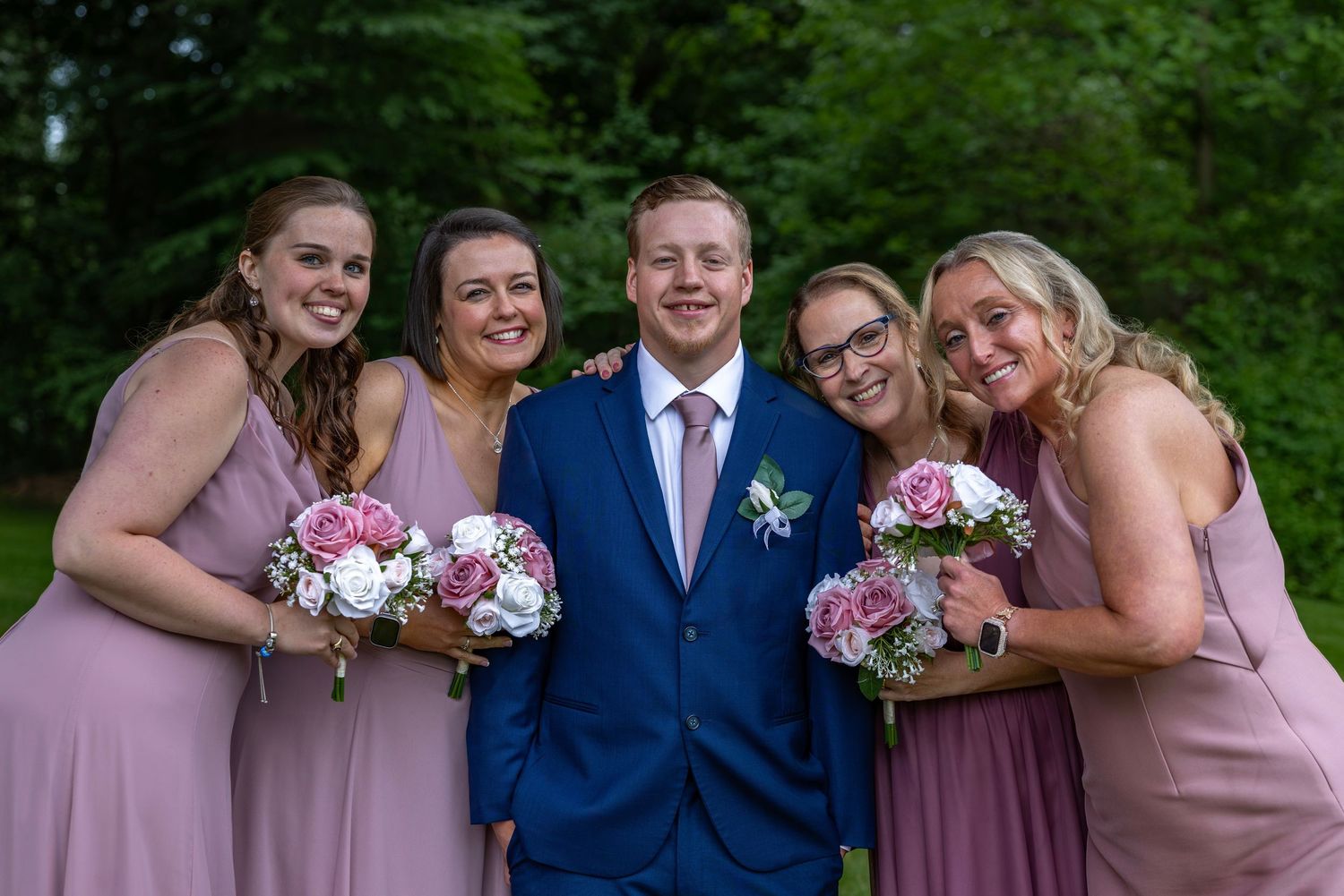 Groom in blue suit poses with four bridesmaids in dusty rose dresses holding bouquets outdoors.