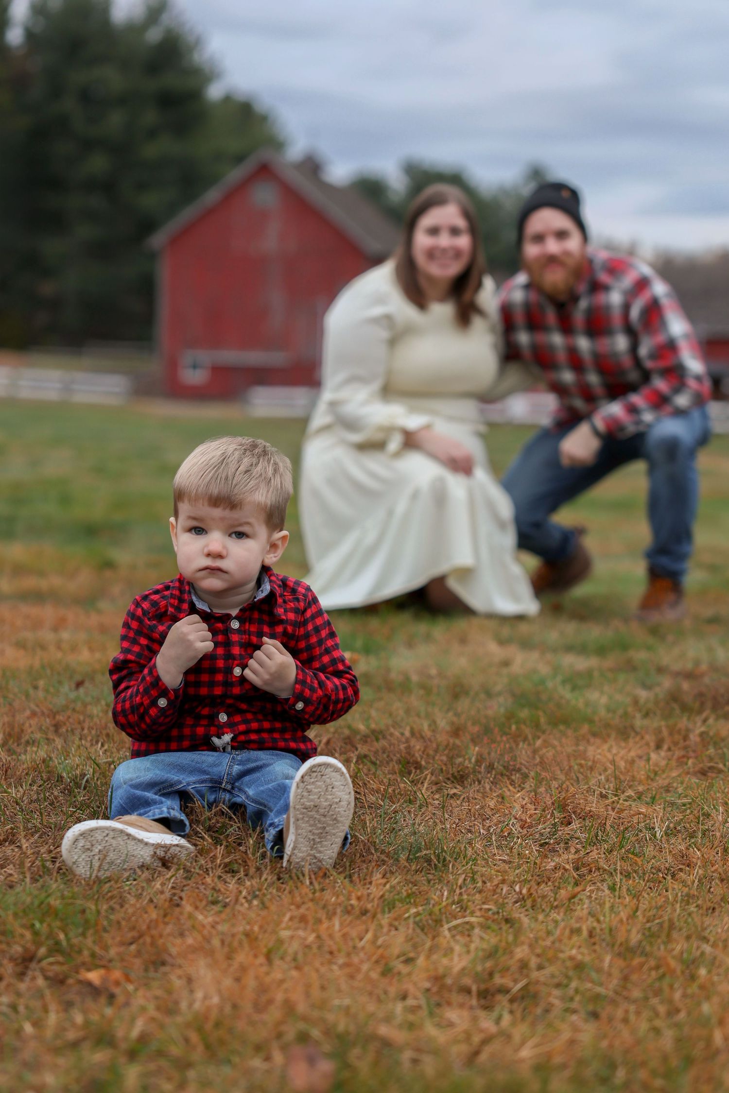 Curious toddler boy in red plaid shirt sitting on grass while parents smile softly in background.