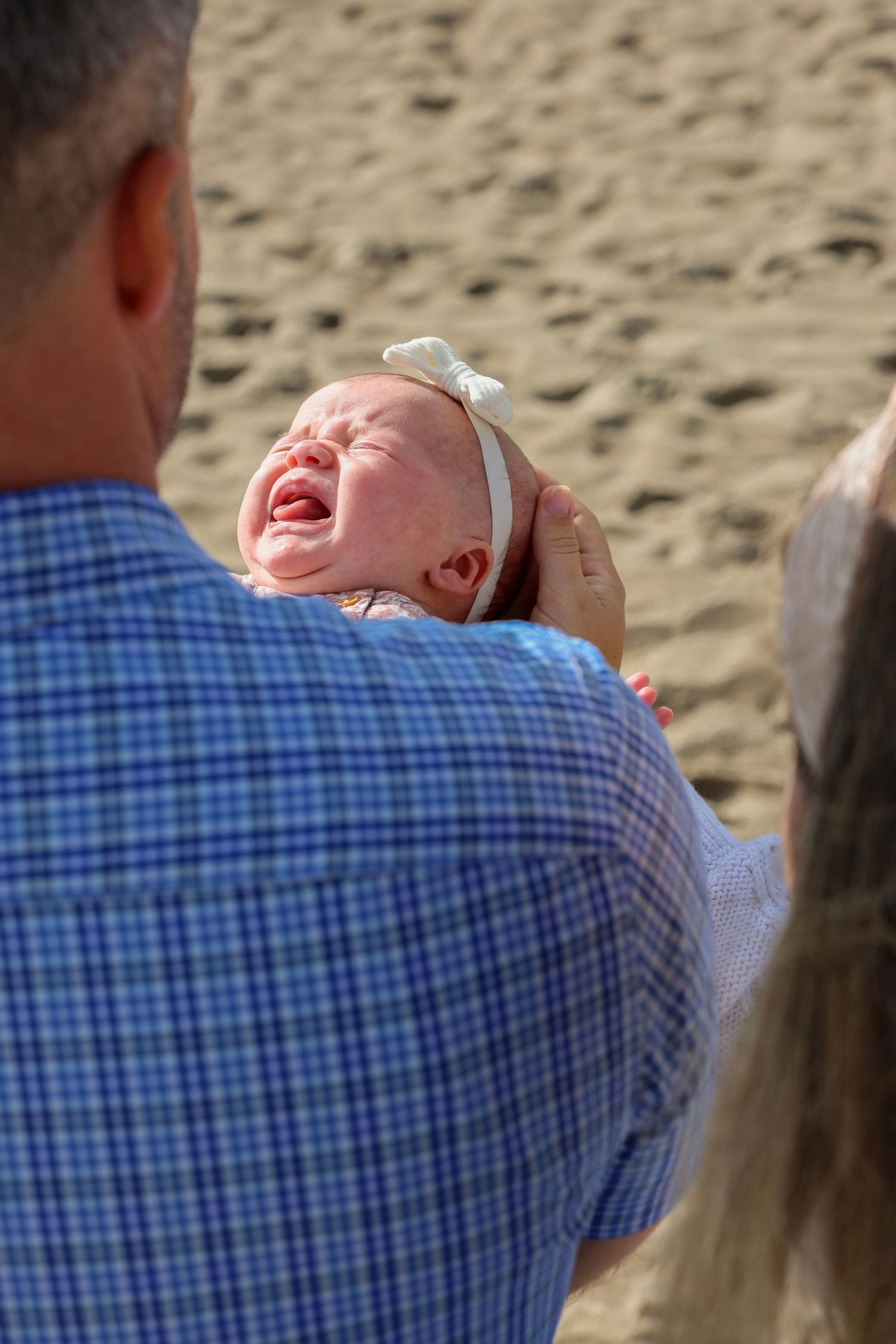 Adult holds crying newborn baby with white bow headband on sandy beach in bright sunlight.