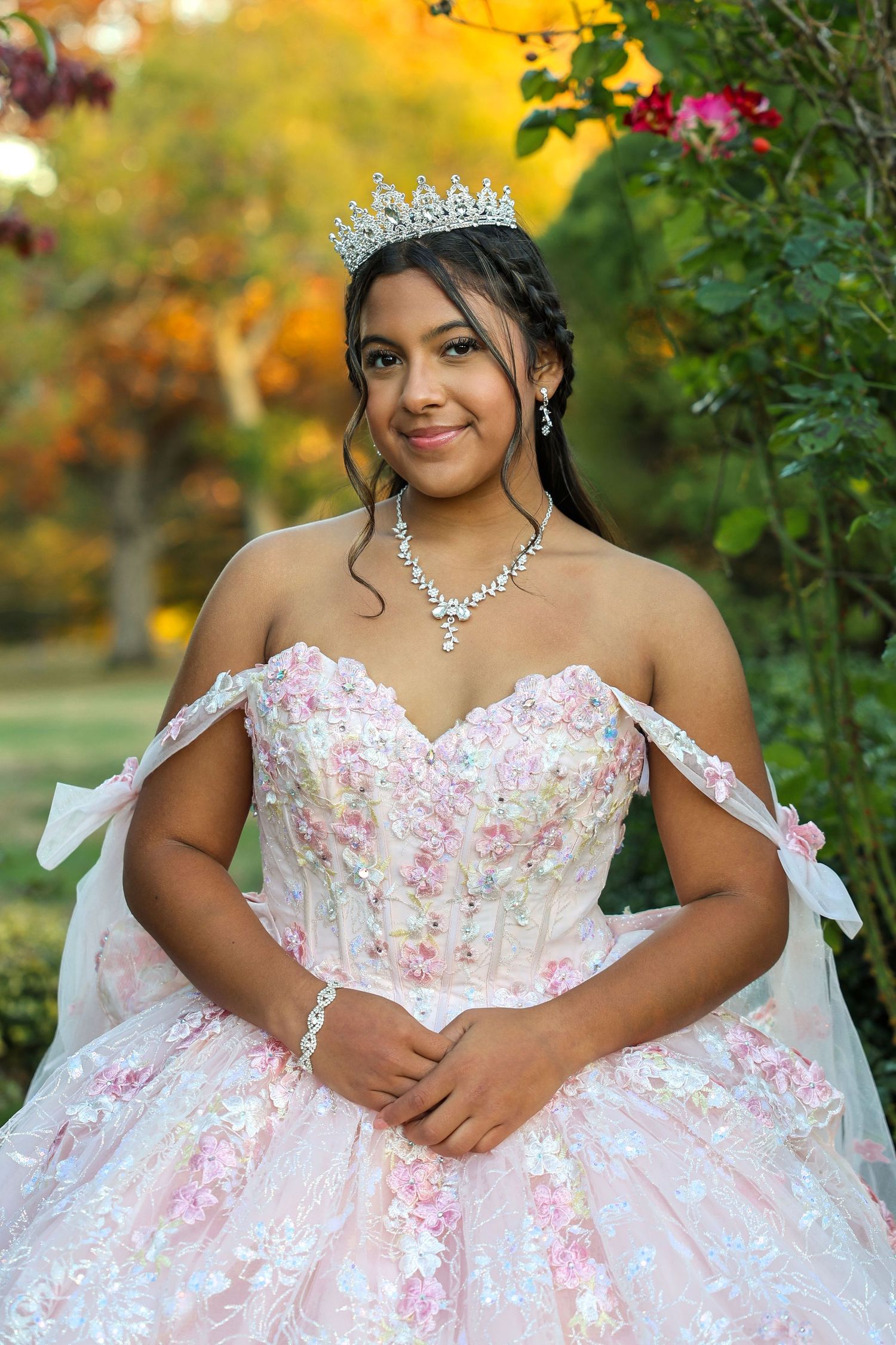 Young Latina girl in a pink floral quinceañera ball gown and diamond tiara posing in autumn garden.