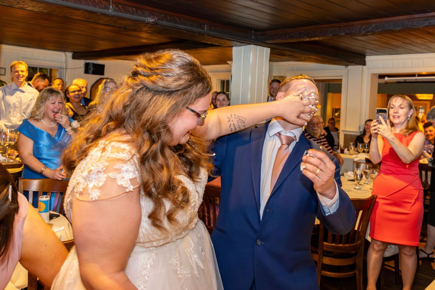 Bride pushes cake into groom's face as wedding guests laugh and photograph the moment.