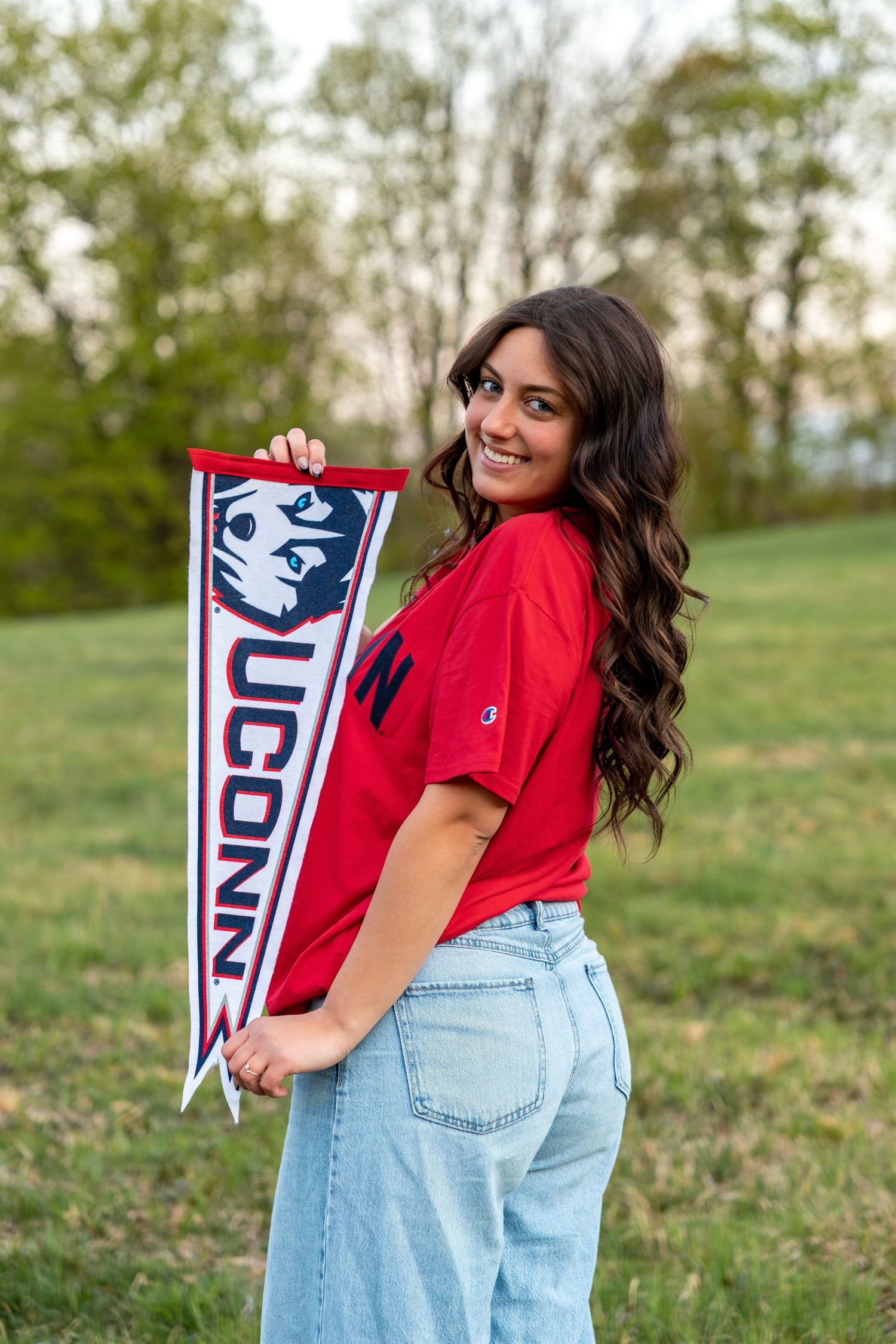 Smiling brunette posing with UConn Huskies pennant flag outdoors in red Champion tee and jeans.