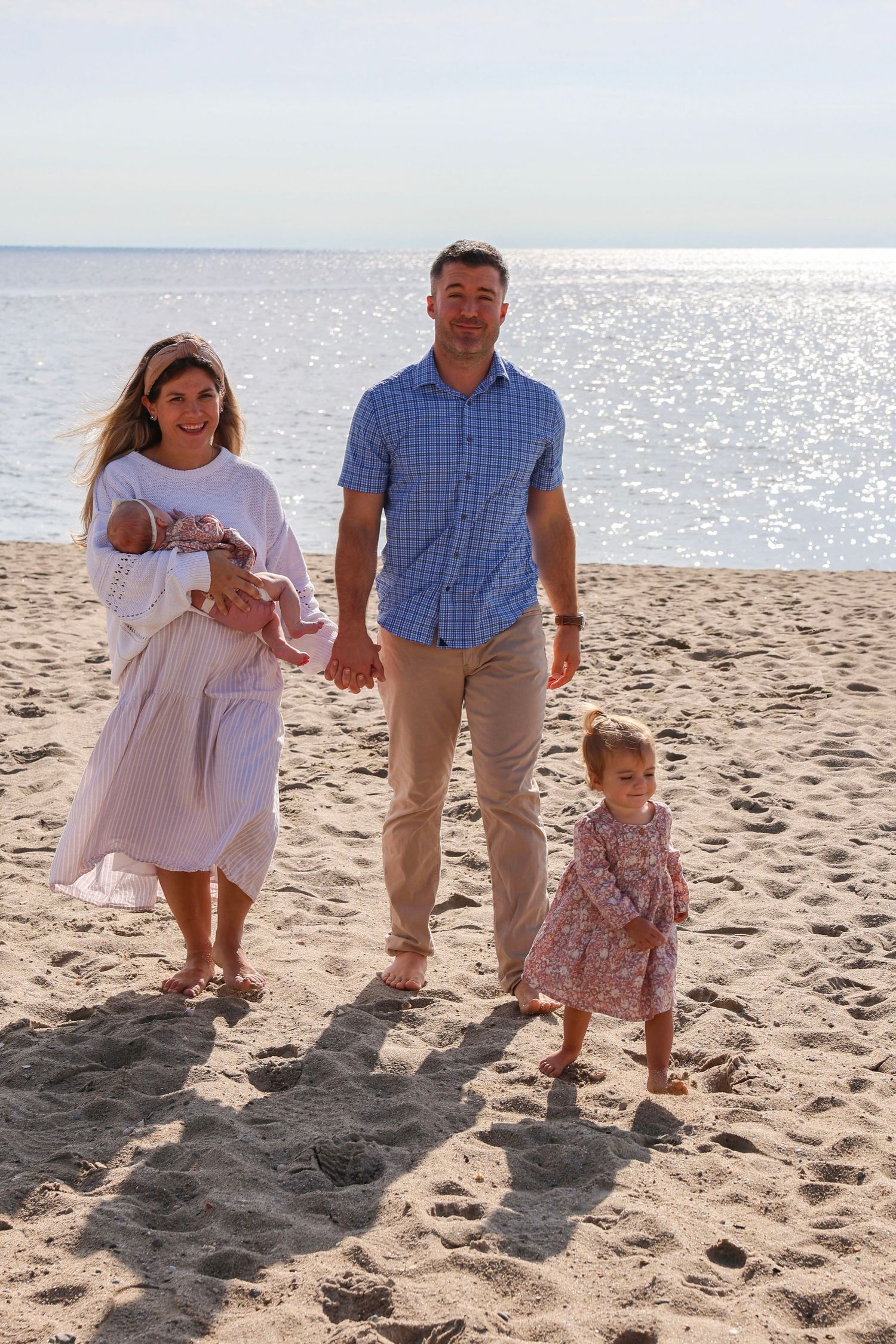 Smiling family with mother holding newborn and father walking with toddler daughter on sunny beach.