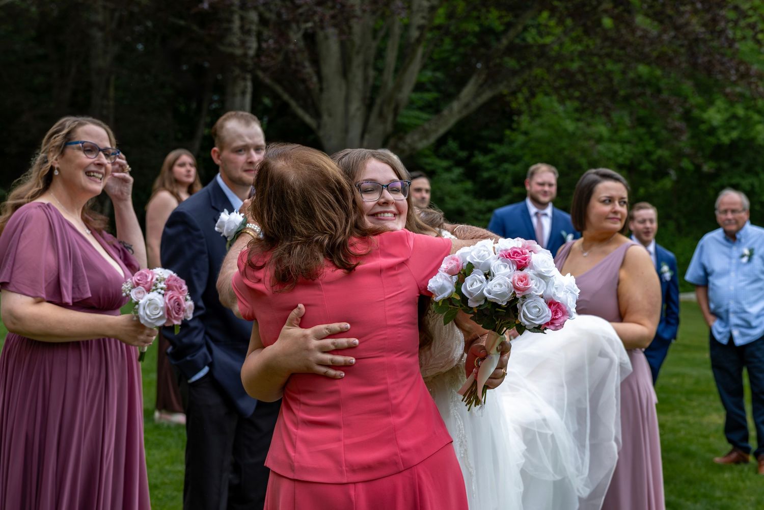 Bride in white gown receives warm hug from guest in coral dress during outdoor wedding celebration.
