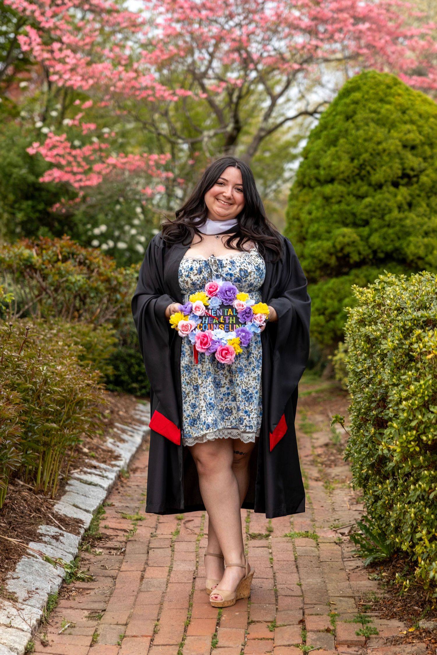 Graduate in black gown walks brick garden path holding flower-decorated cap amid pink blossoming spring trees.