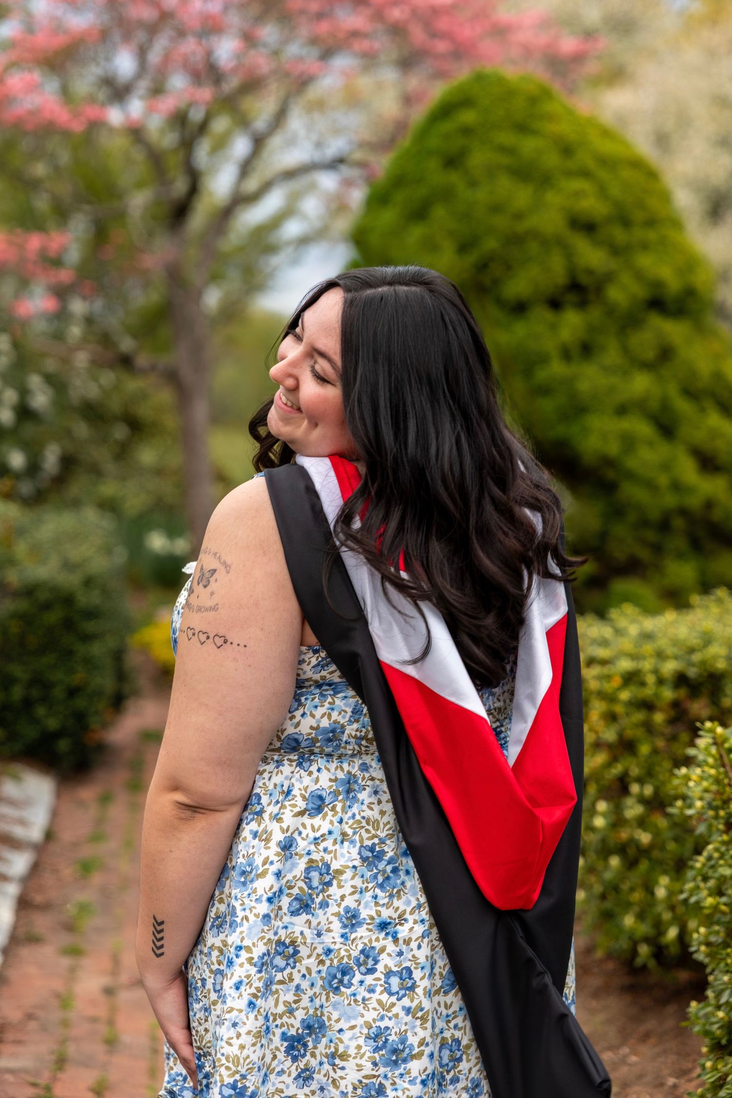 Laughing graduate in floral dress with black and red academic gown looks over shoulder on brick garden path.