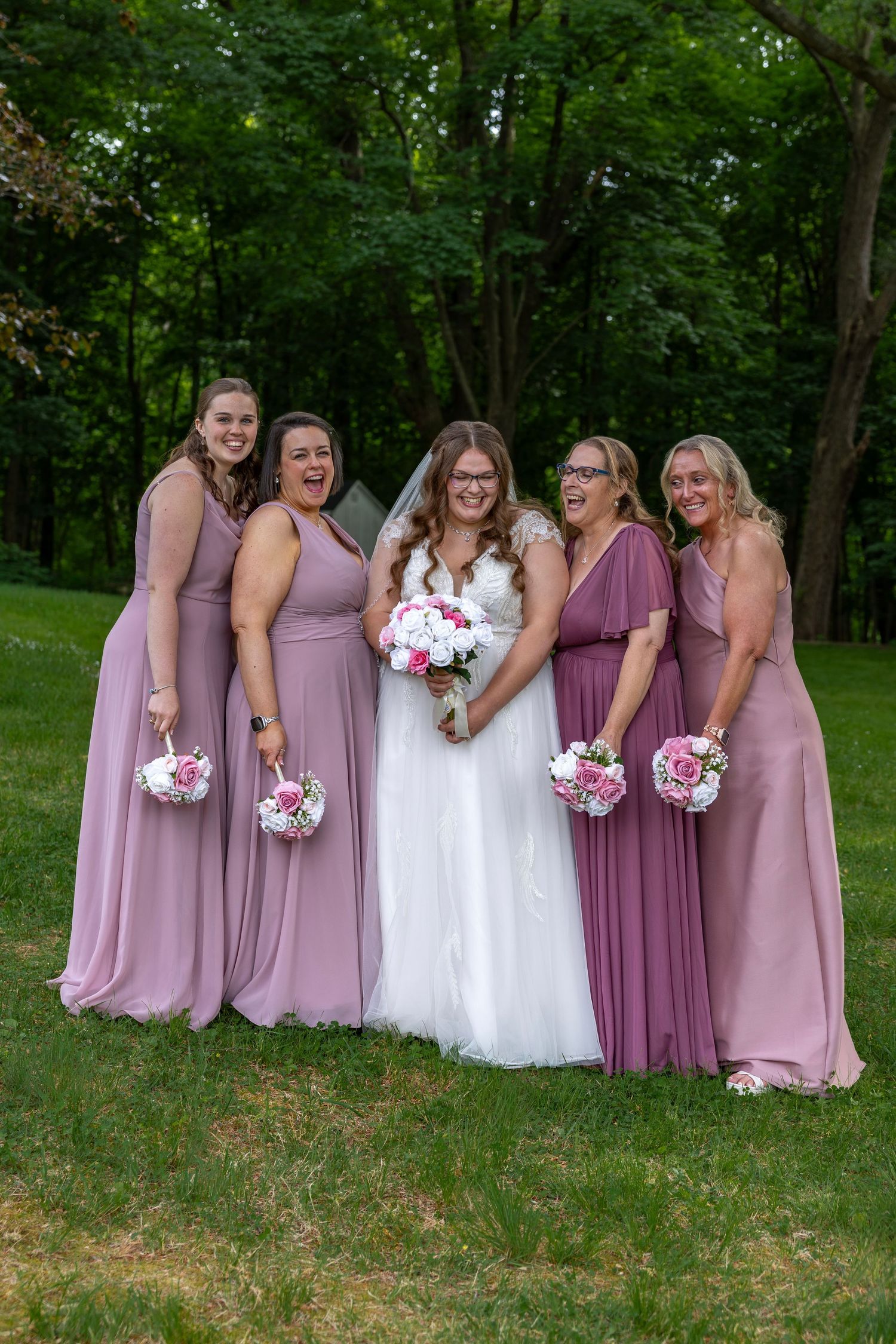 Bride with four bridesmaids in mismatched mauve gowns laughing together in outdoor wedding party photo.