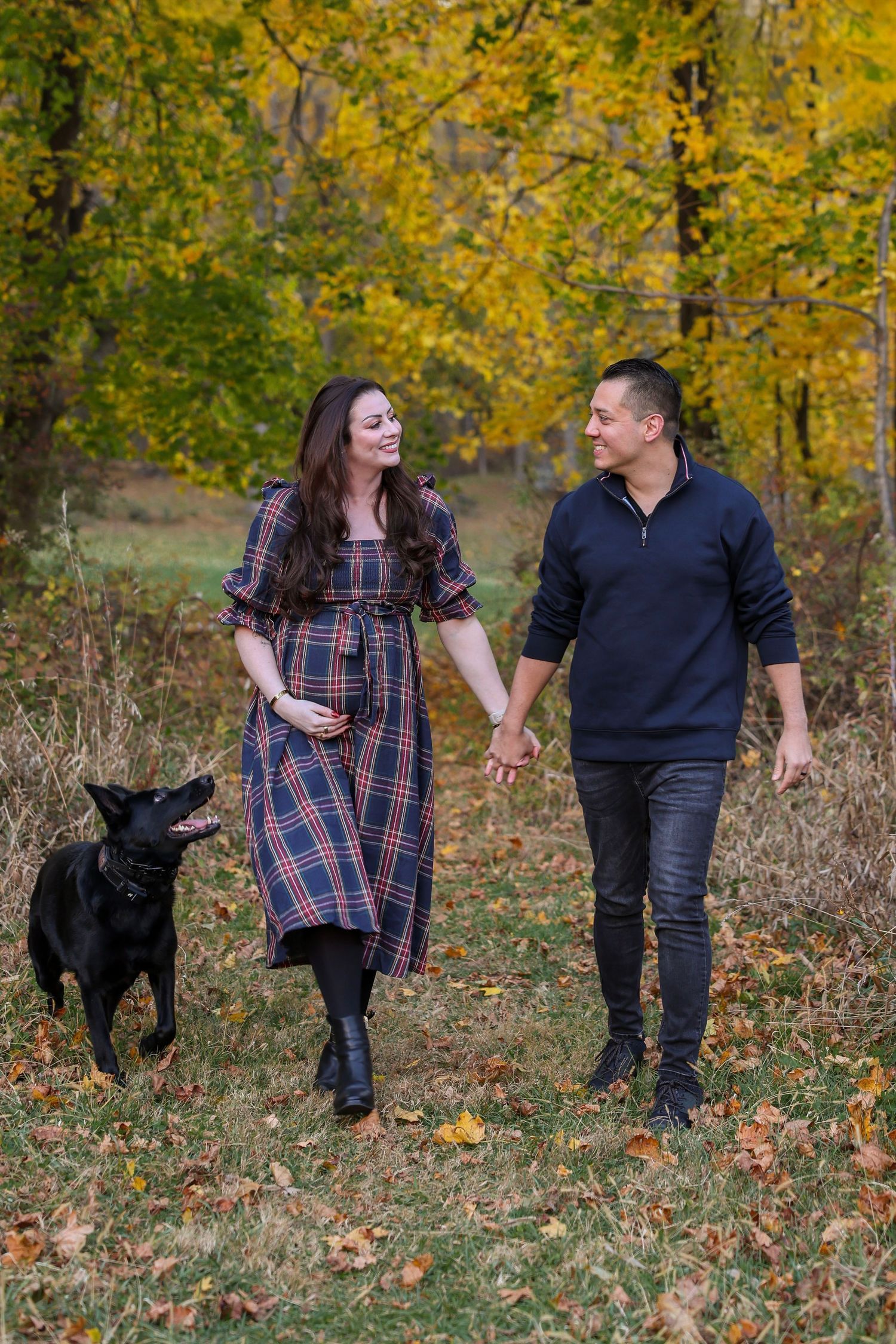 Pregnant couple holds hands walking with black dog among vibrant autumn foliage.