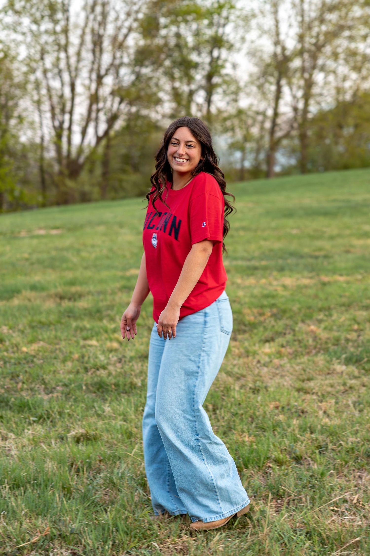 Laughing young woman in red UConn Champion tee and wide-leg jeans standing in outdoor grassy field.