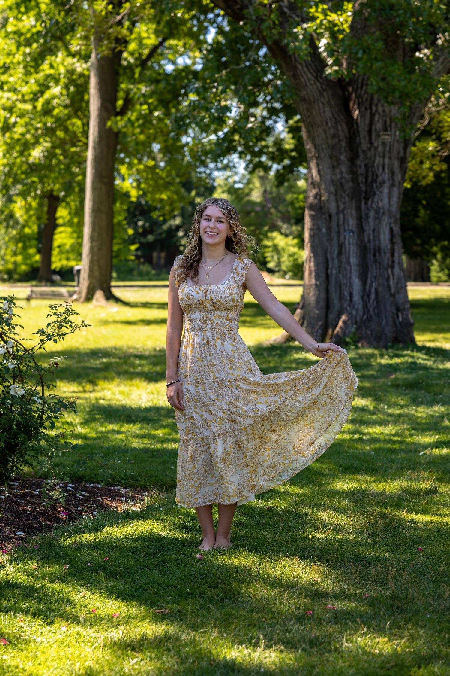 Young woman in floral midi dress twirling barefoot in sunlit green park surrounded by trees.