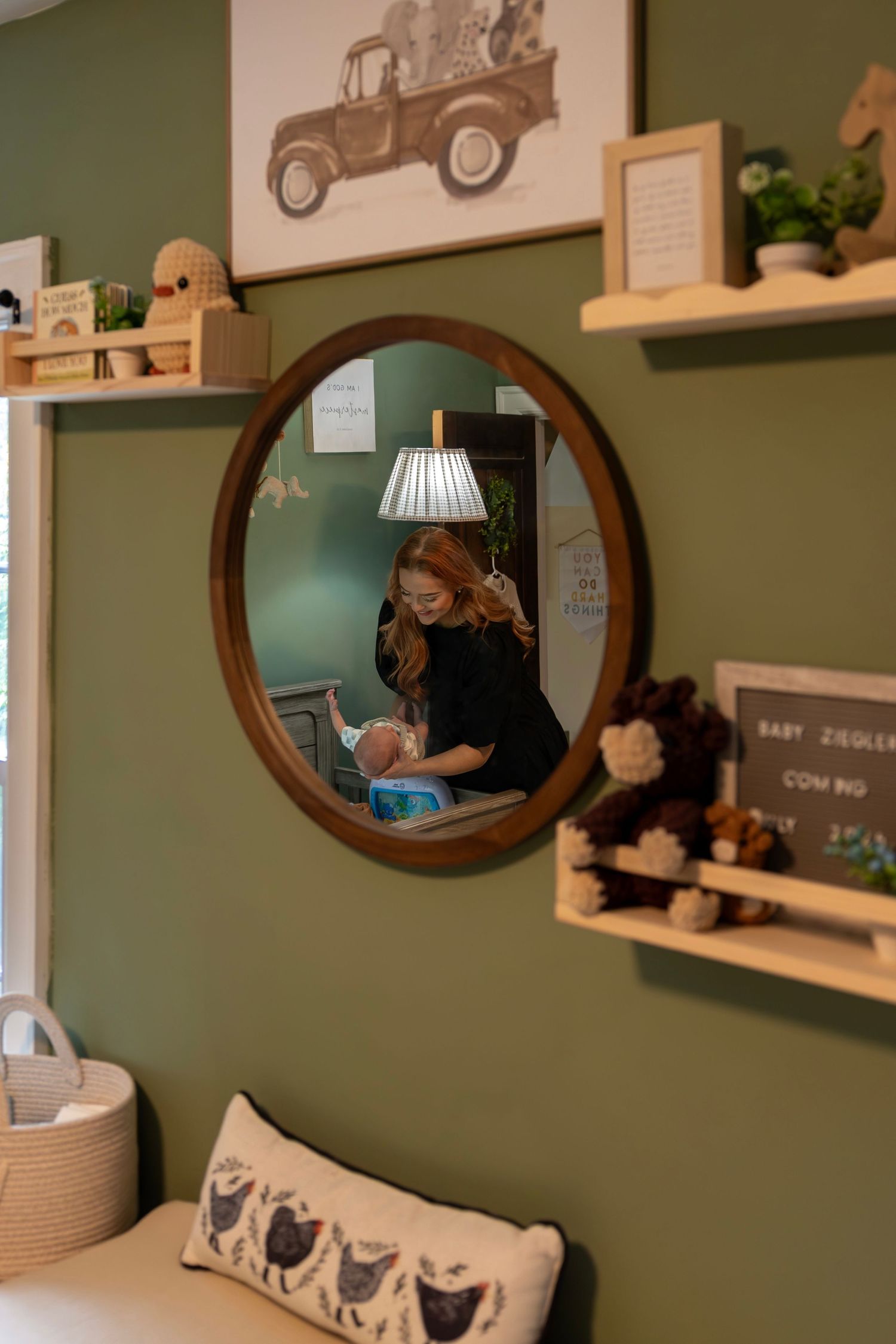 A mother tends to her newborn in a green nursery, reflected in a round wooden mirror surrounded by rustic shelves.