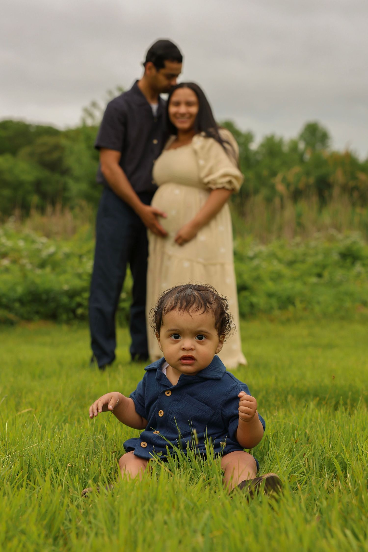 Curious baby in blue romper sits in grass with expecting parents blurred behind.