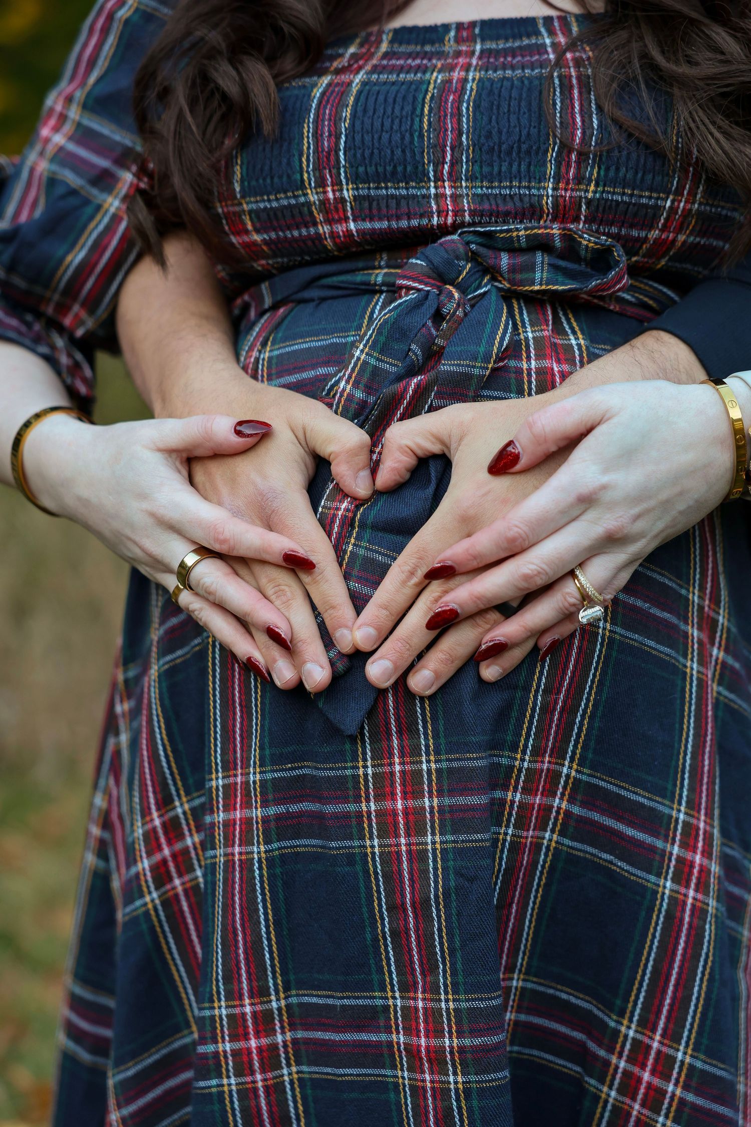 Couple forms heart shape with hands on pregnant belly wearing plaid maternity dress.