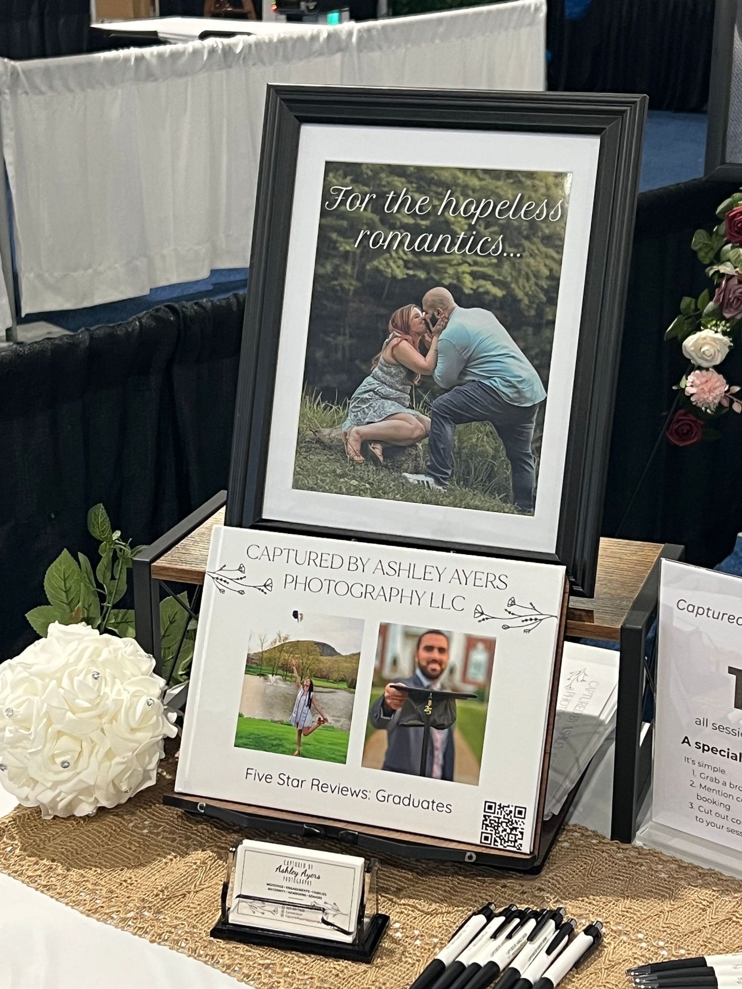 A framed romantic couples photo with hopeless romantics quote and five-star graduate reviews at a photography expo booth display.