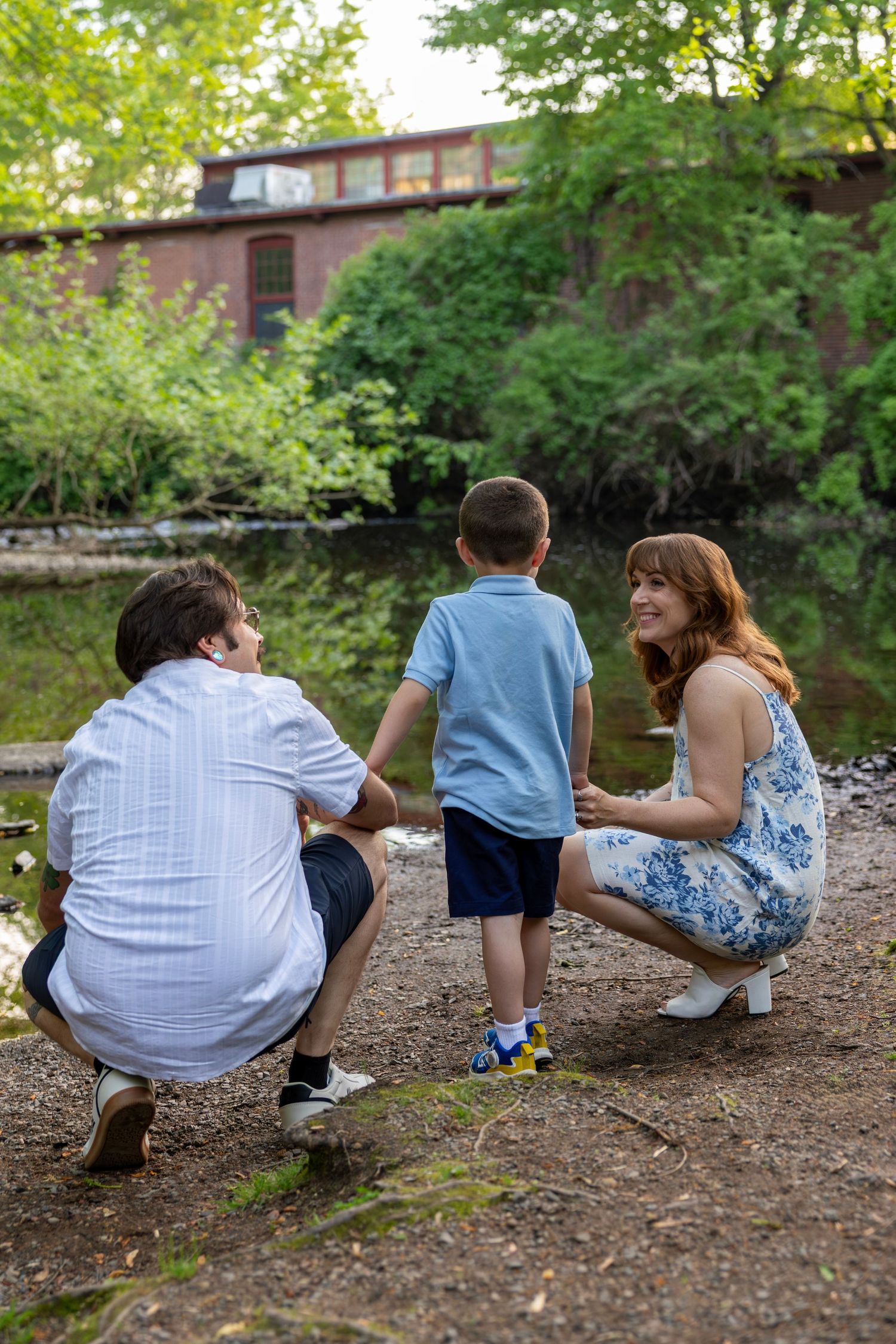 Family of three crouches together by a serene river with a red brick building visible in the background.