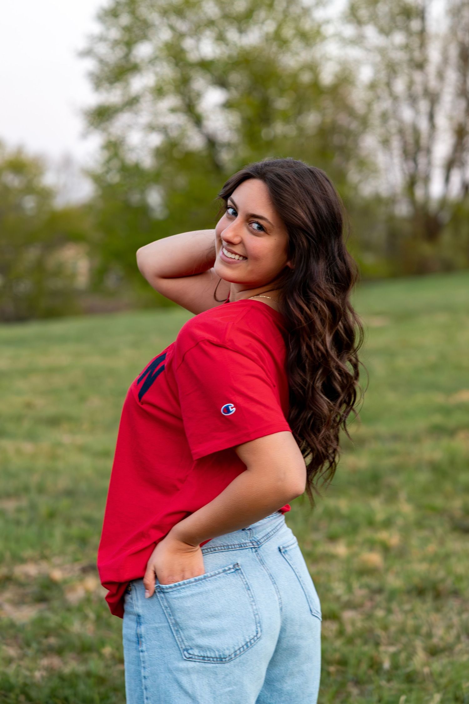 Smiling brunette woman glancing back over shoulder in red Champion tee and jeans in grassy field.