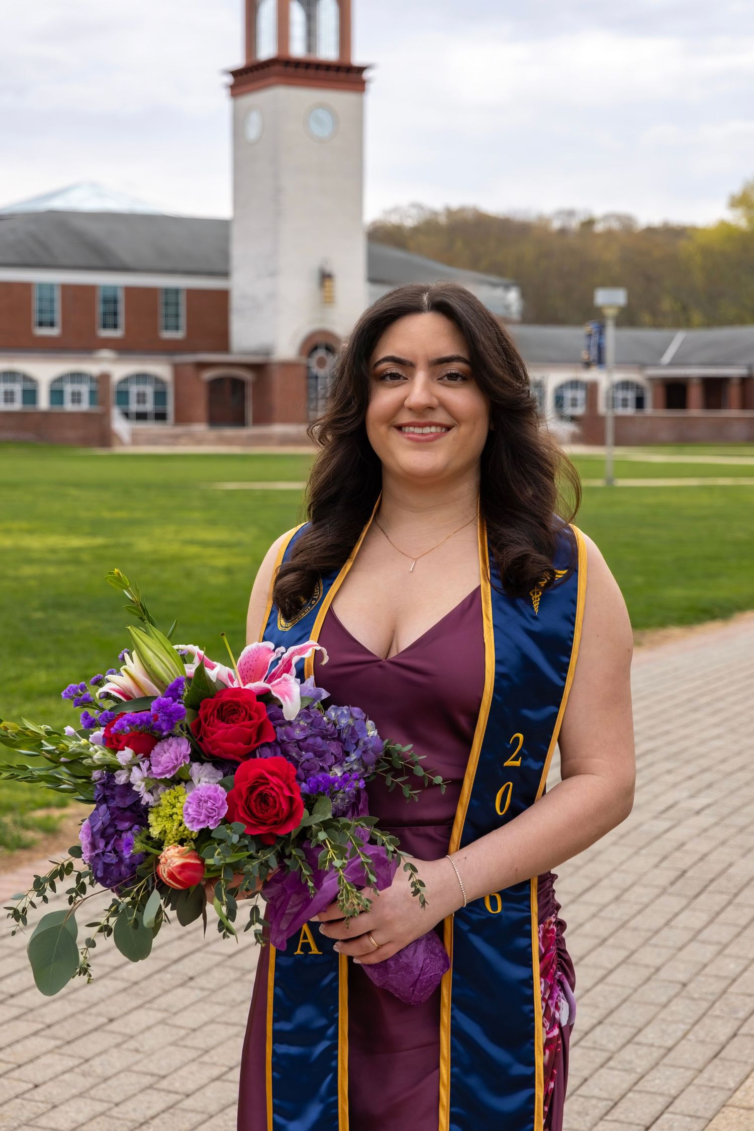 Smiling female graduate in purple sash holding colorful bouquet near brick campus building.