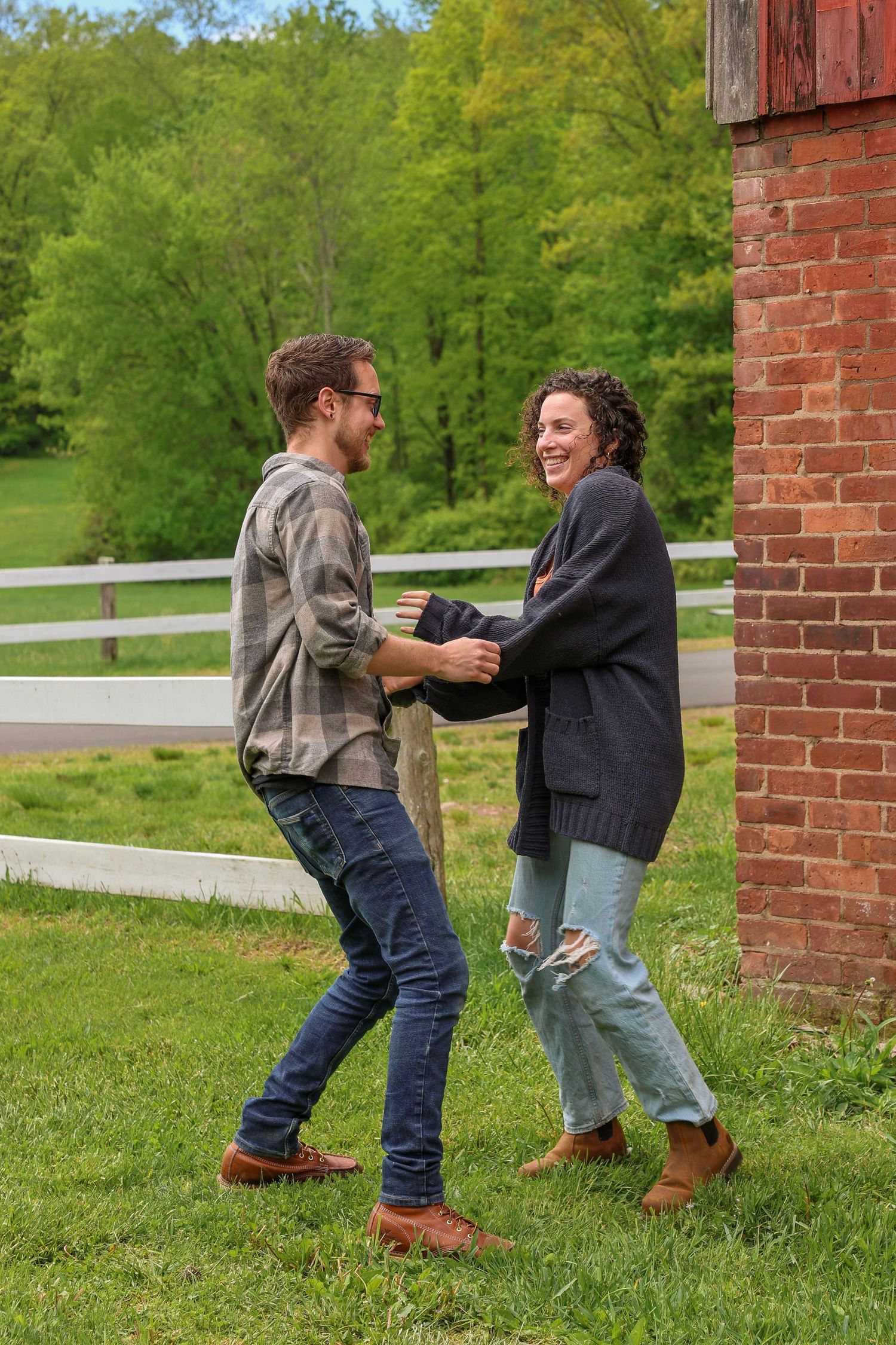 A smiling couple dancing and laughing together on green grass beside a rustic red brick barn.