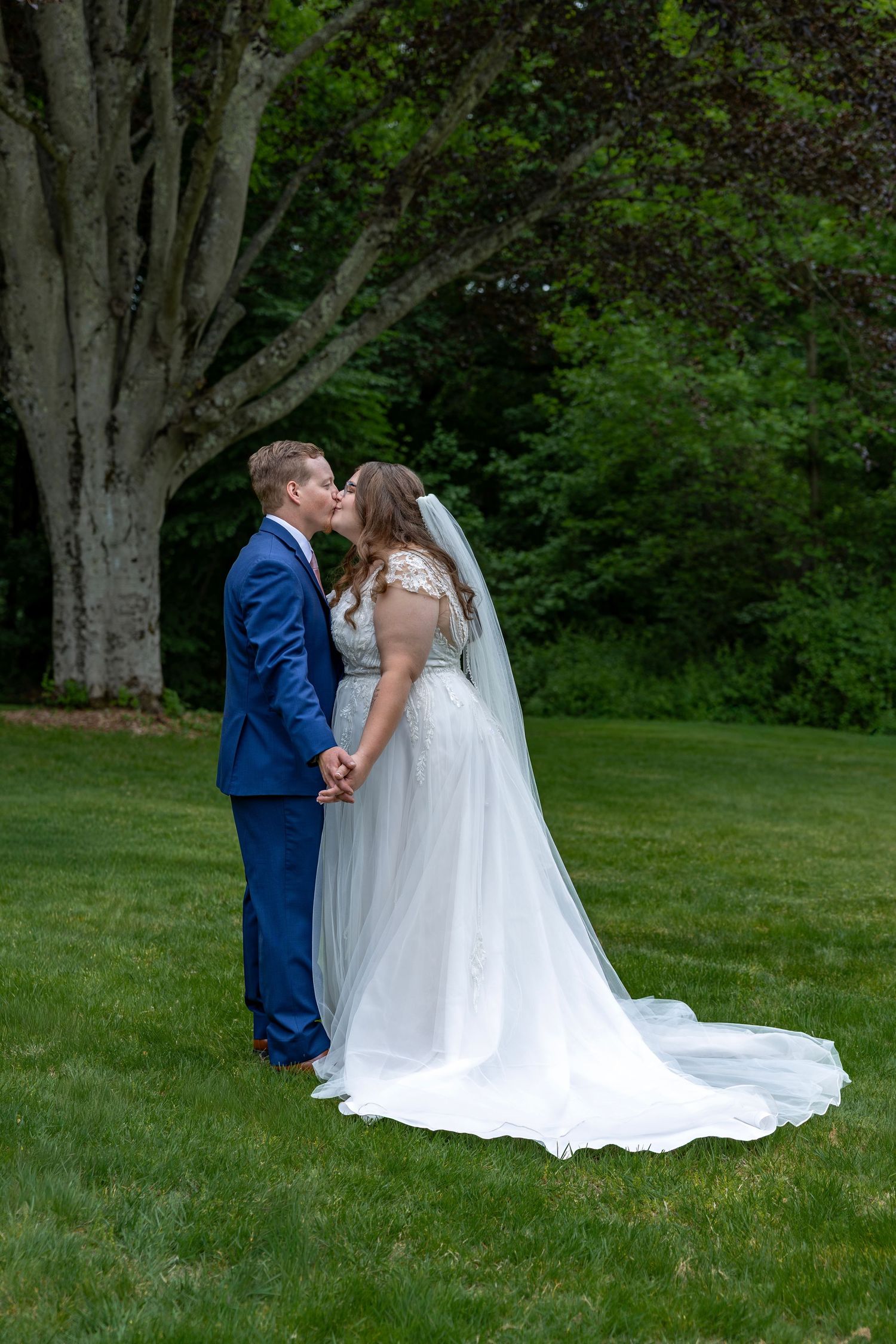 Bride and groom share romantic kiss on green lawn with large tree backdrop at wedding.