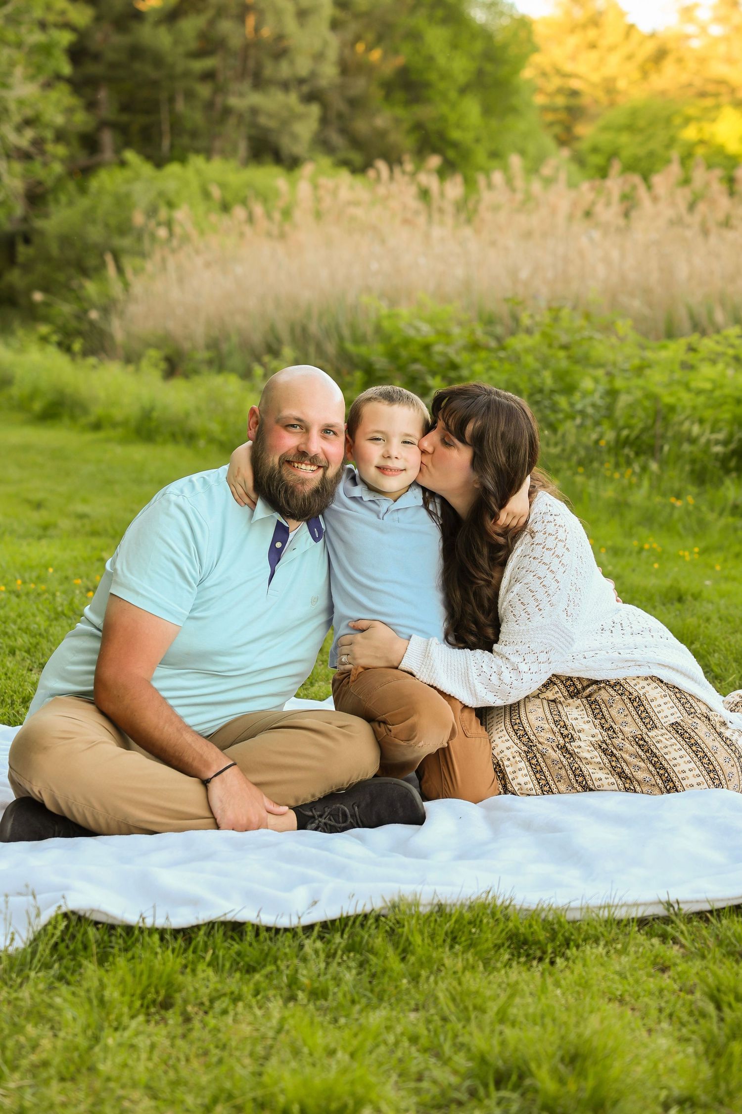 A mother kisses her son on the cheek as the family sits together smiling on a blanket outdoors.