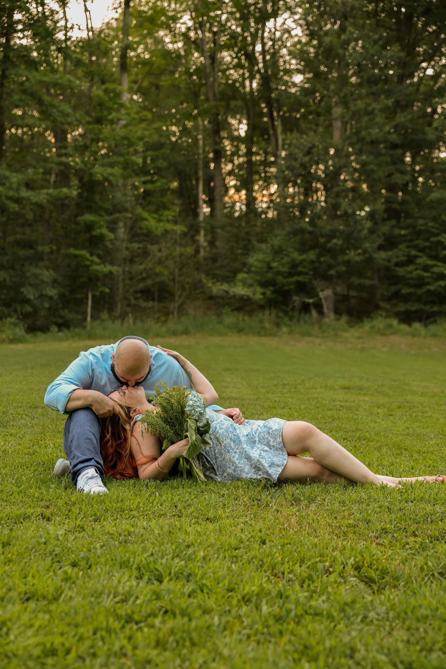 Romantic couple sharing a kiss on green grass, woman reclining with a wildflower bouquet in a park setting.