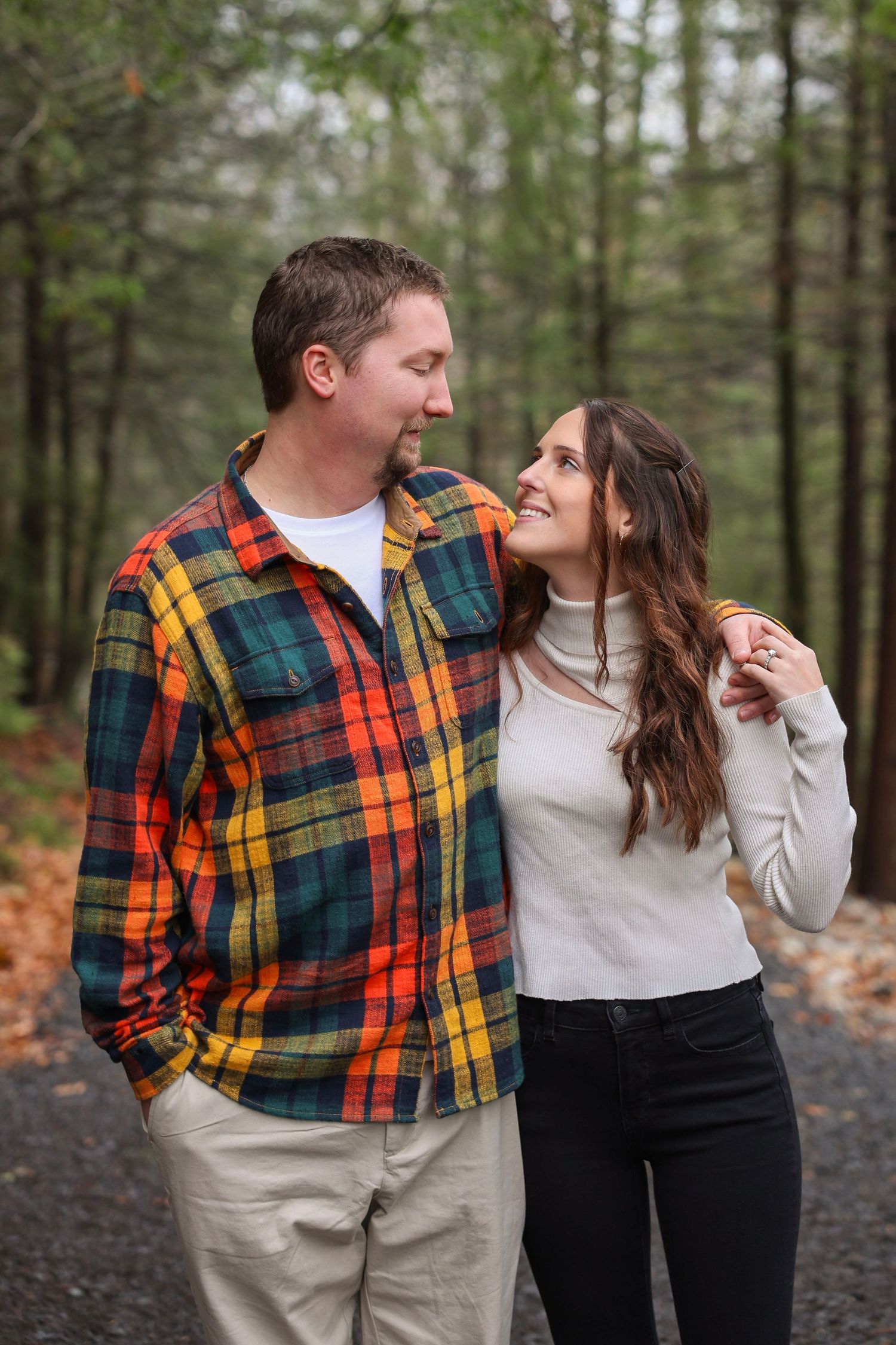 A couple walks closely together on a forest path, smiling at each other in a colorful plaid shirt.