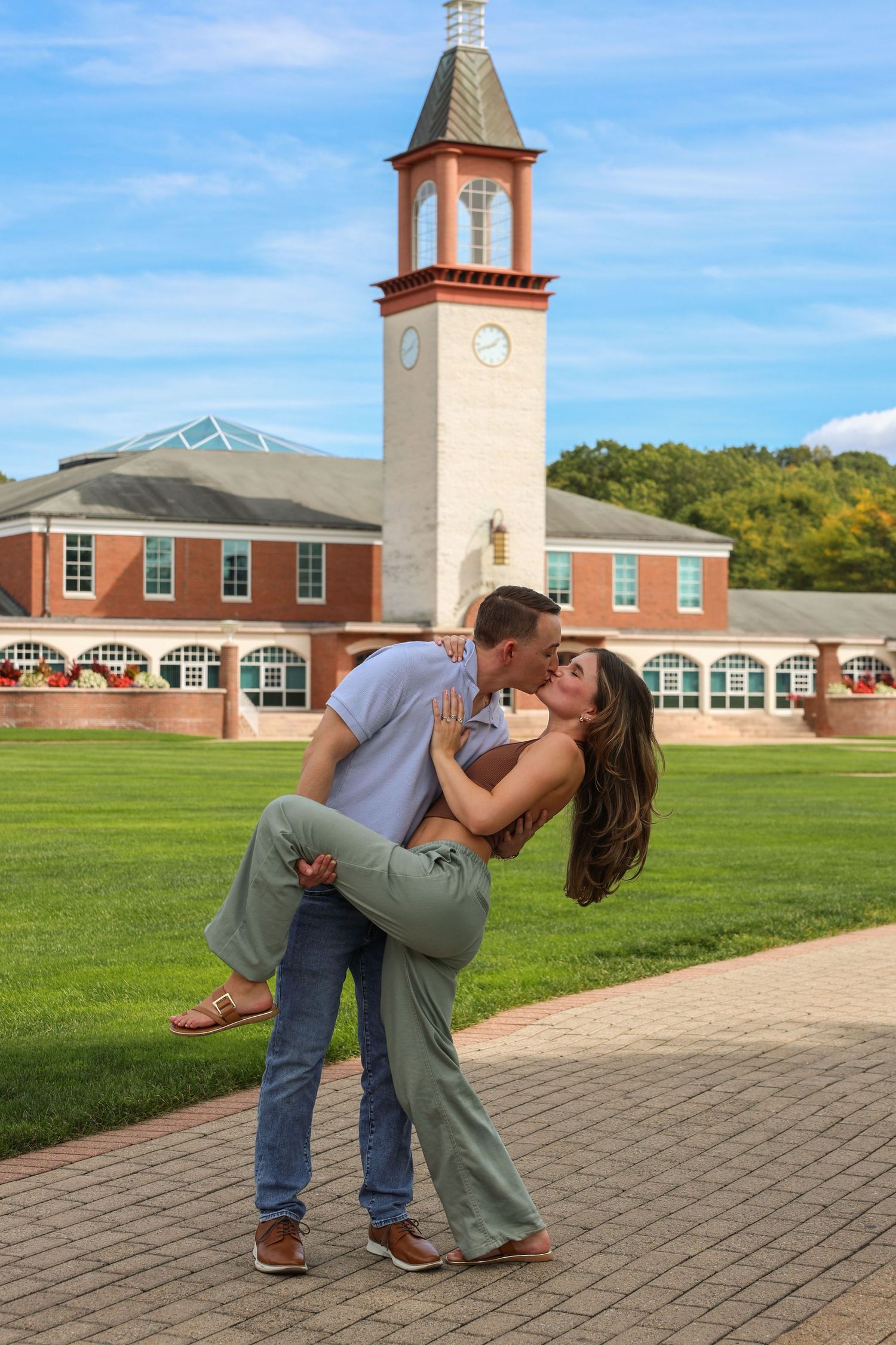 Man dipping and kissing his fiance in a romantic pose on a brick pathway with a clock tower behind them.