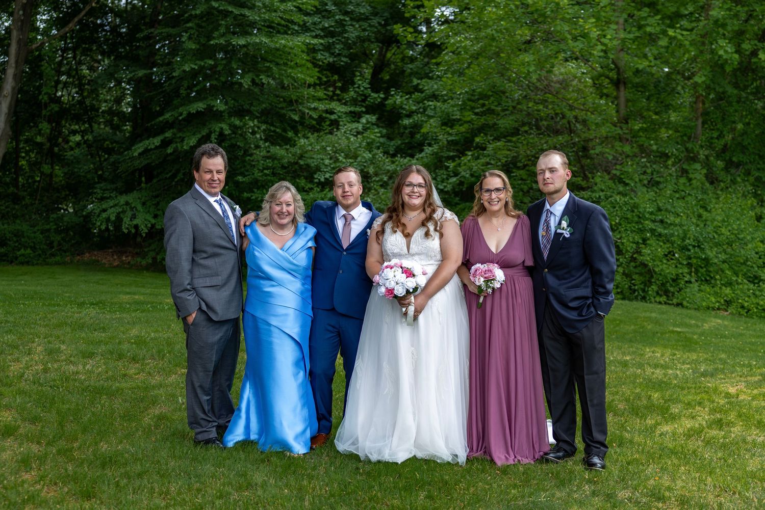 Bride and groom pose with four family members on green lawn, bride holding pink and white bouquet.