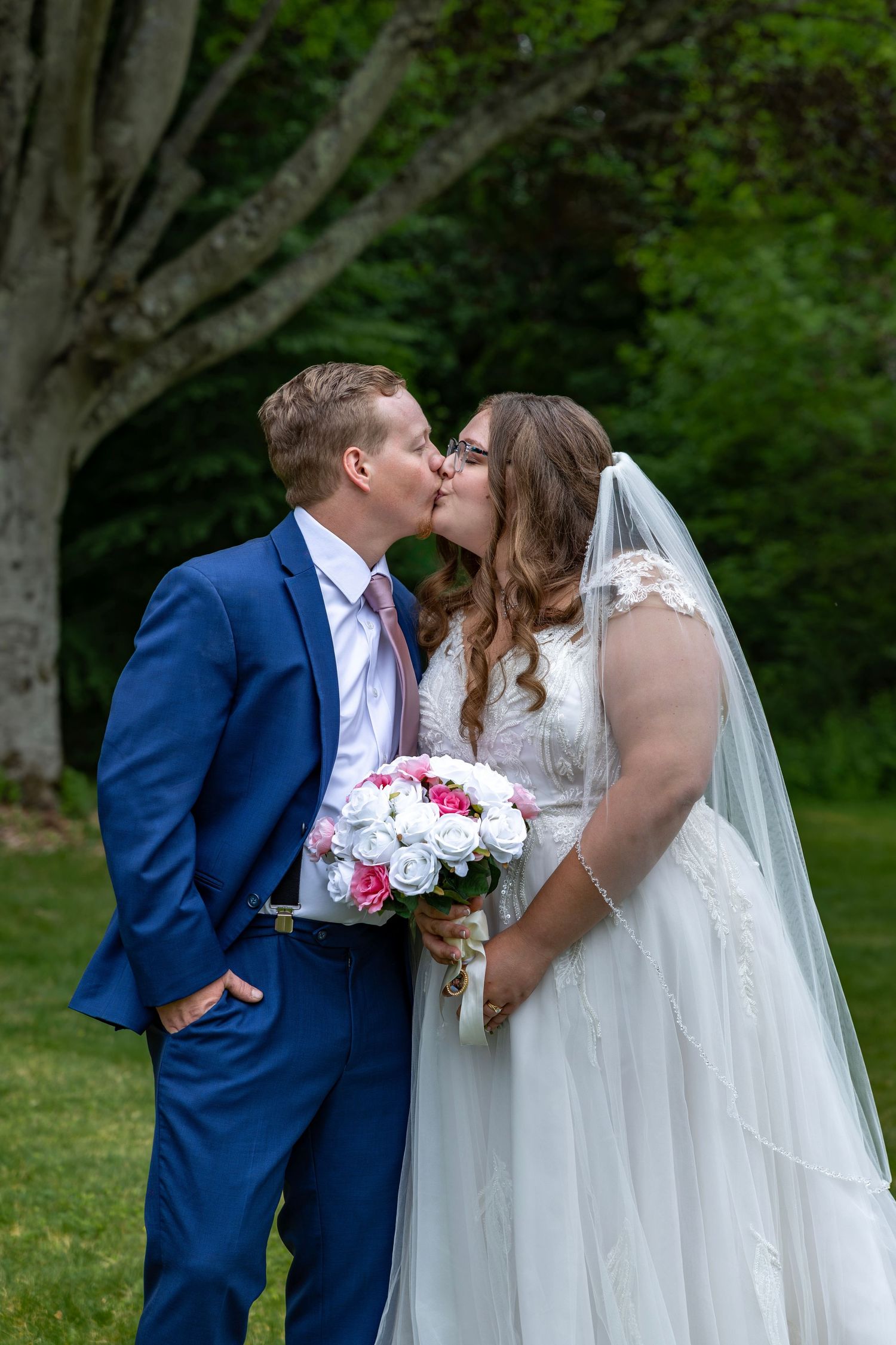 Newlyweds kissing outdoors as bride holds pink and white bouquet, groom in blue suit with pink tie.