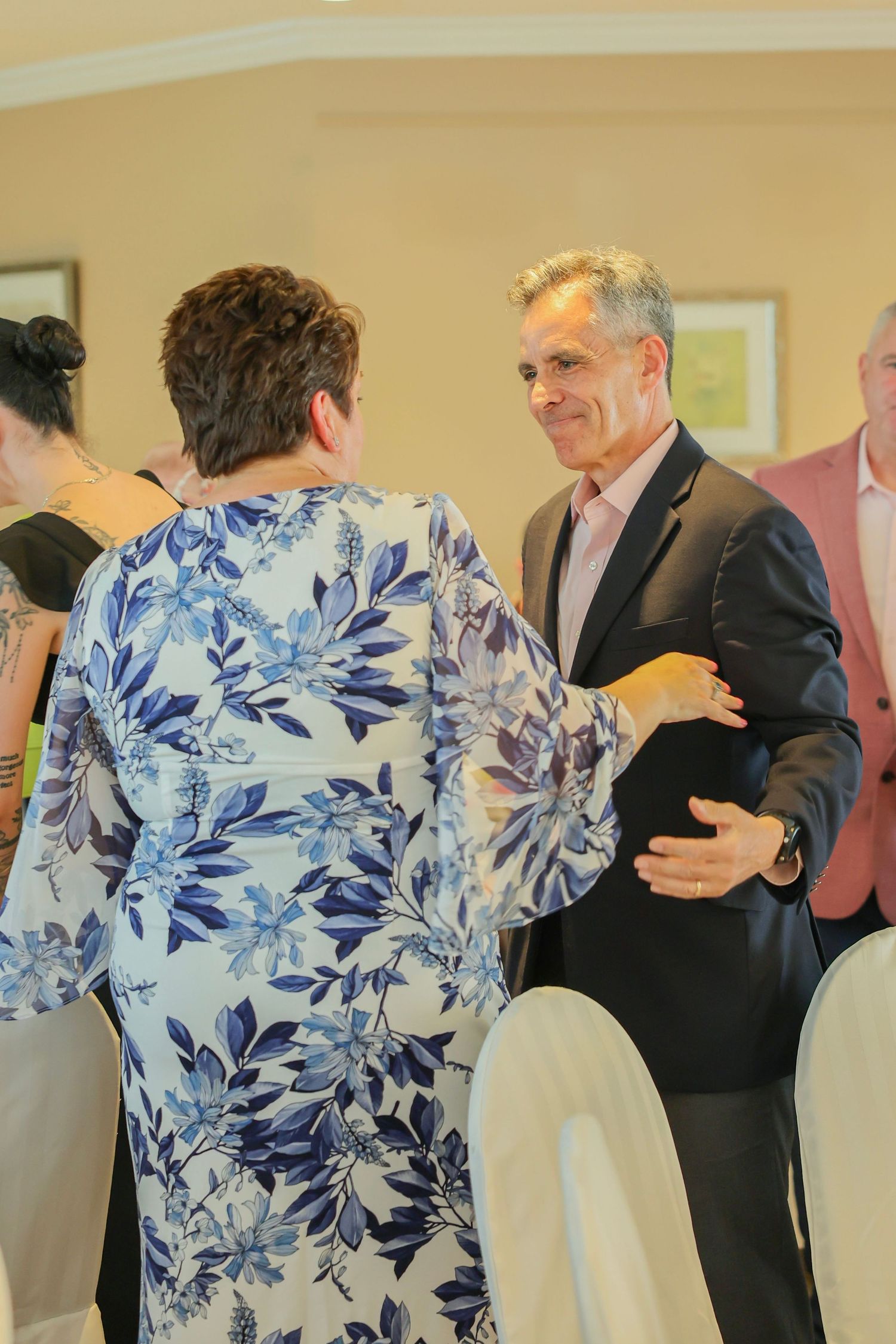 Man in dark blazer warmly greeting woman in blue floral dress at an indoor social gathering.