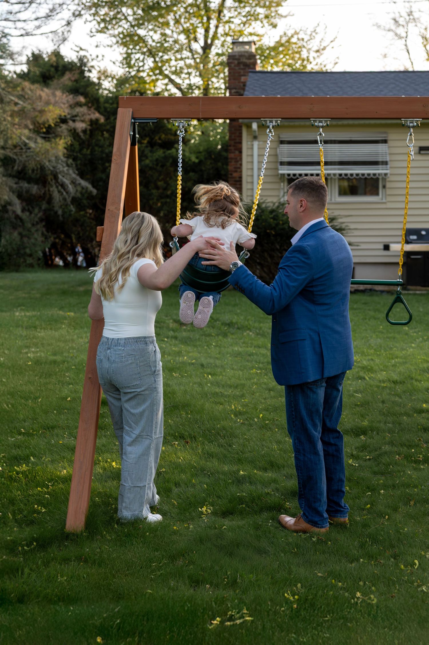 Family of three playing at backyard swing set during golden hour, parents lifting toddler near wooden swing frame.